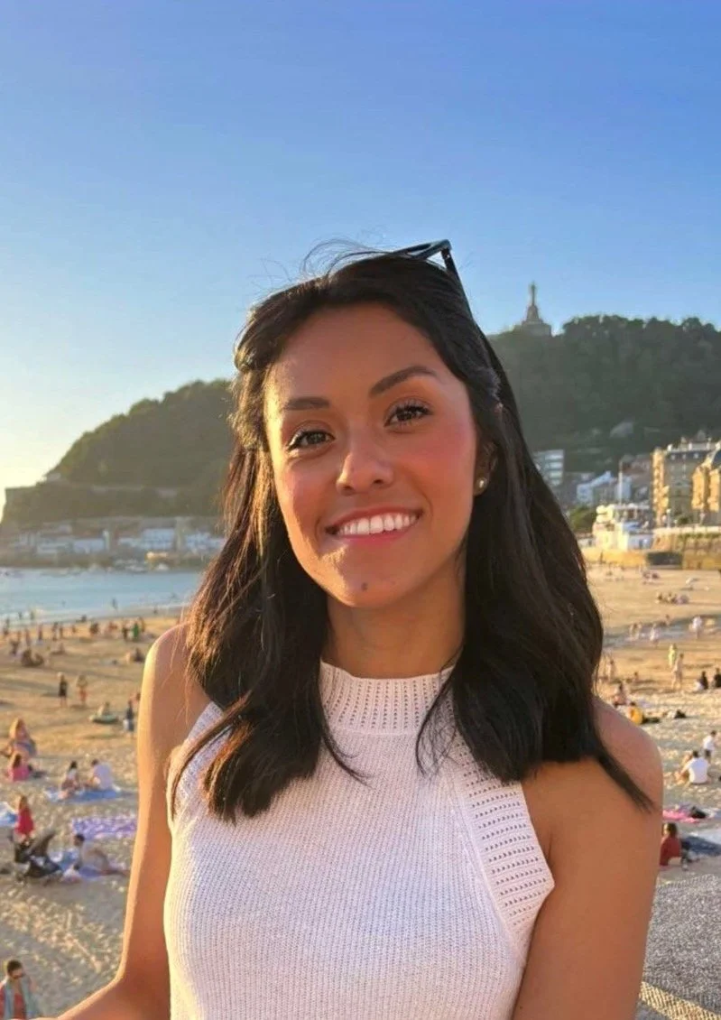 Jeune femme souriante avec des cheveux noirs et une robe blanche, posant sur la plage avec la ville et une colline en arrière-plan, soleil couchant.