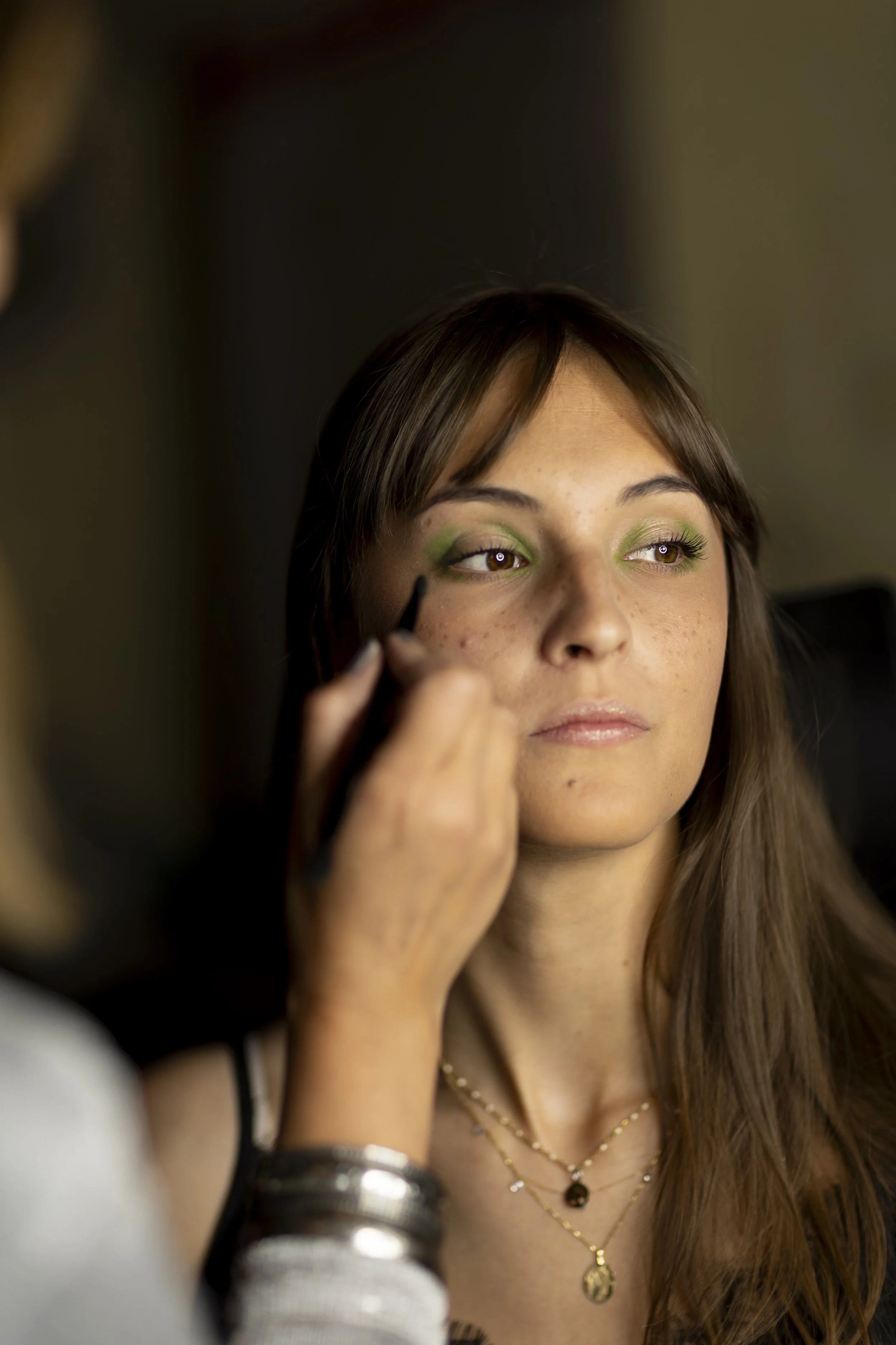 Une femme avec des yeux maquillés en vert, recevant un maquillage de l'assistante, portant des colliers dorés, dans un intérieur éclairé.