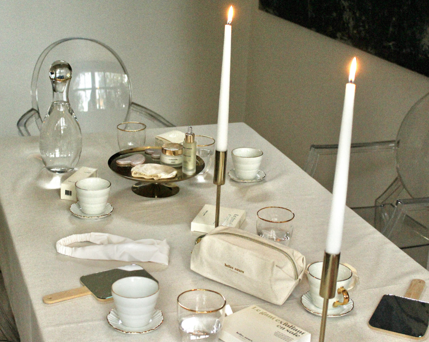 Une table élégante ornée de chandelles allumées, assiettes en porcelaine blanche avec des bords dorés, verres à eau, tasses, un plateau doré avec des cosmétiques, un sac beige et divers objets de décoration dans un cadre moderne.