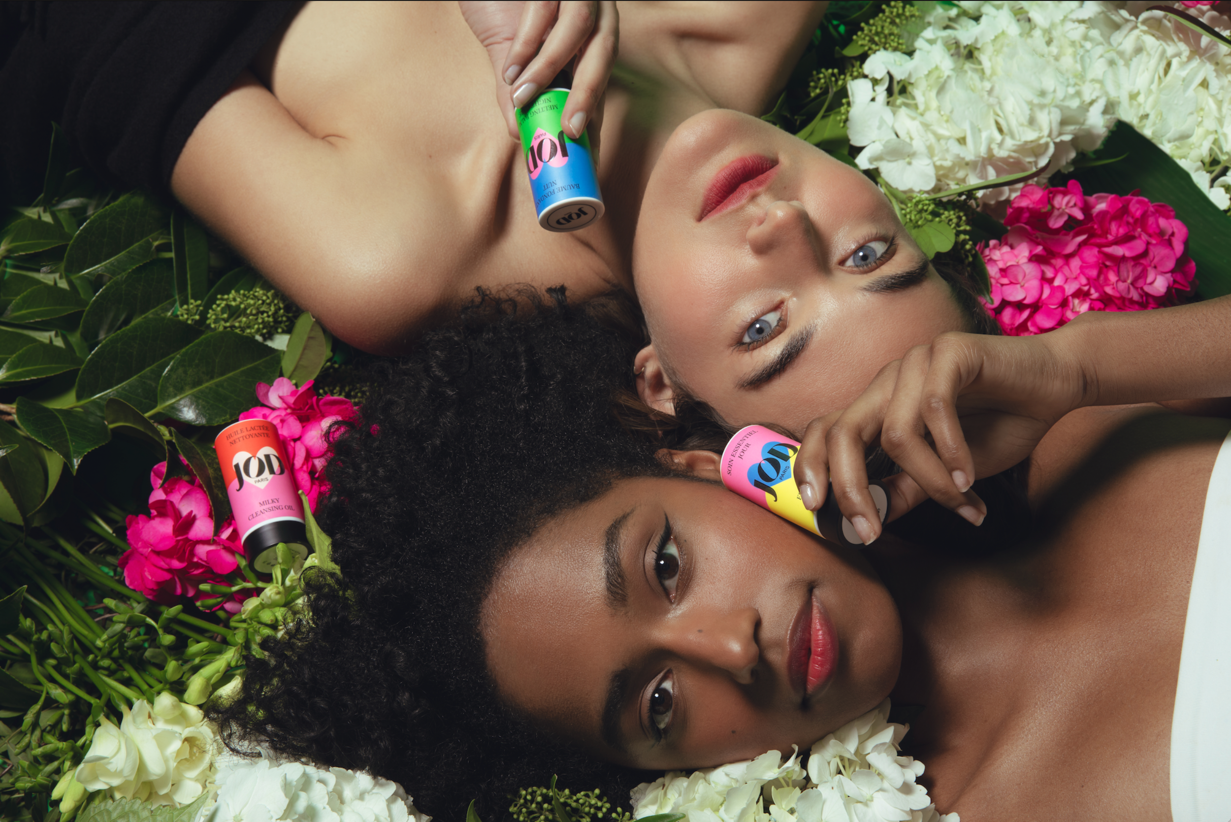 Deux femmes allongées sur un lit de fleurs, tenant des vapoirs colorés au hasard, entourées de fleurs blanches et roses, avec un maquillage naturel et des expressions sereines.