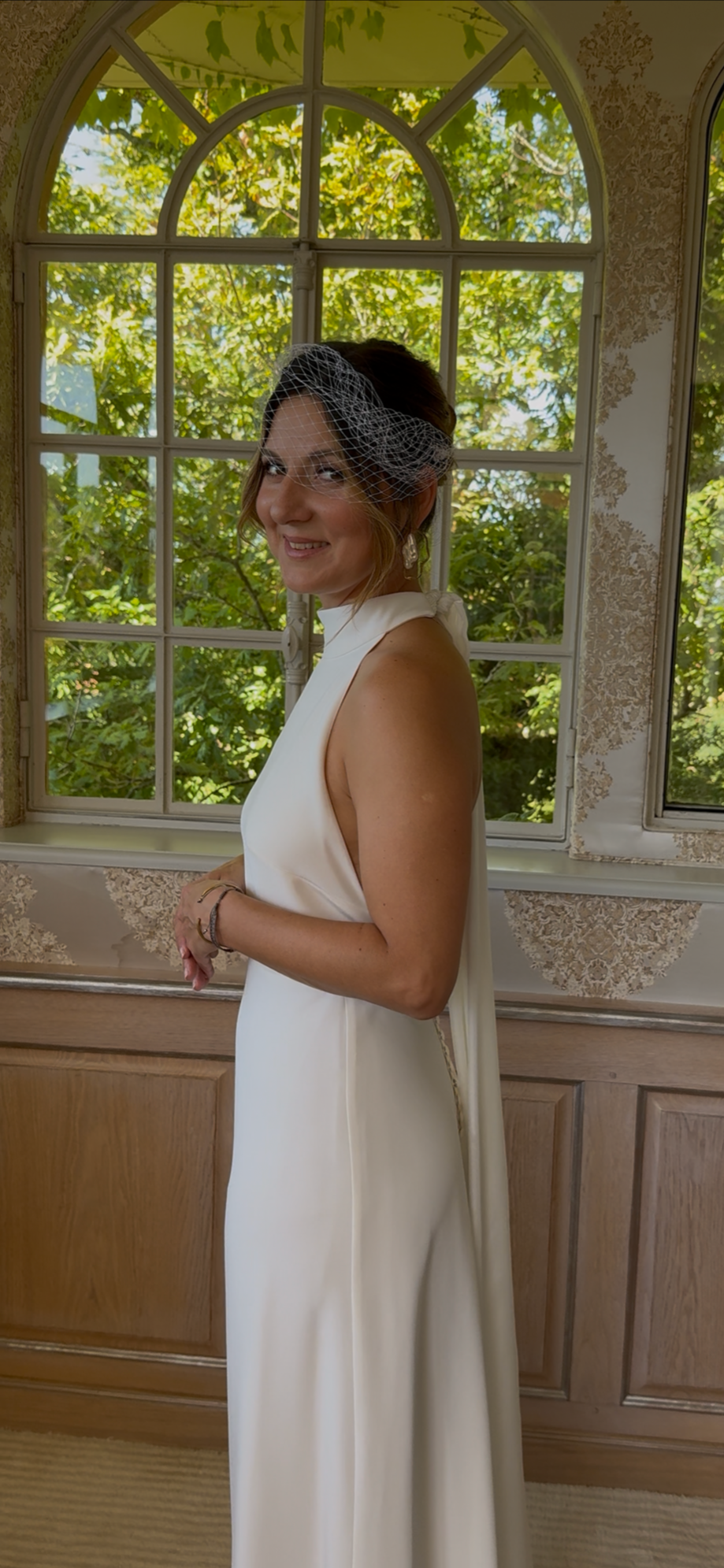 Une femme en robe blanche et voile de mariée, debout près d'une grande fenêtre avec vue sur des arbres verts.