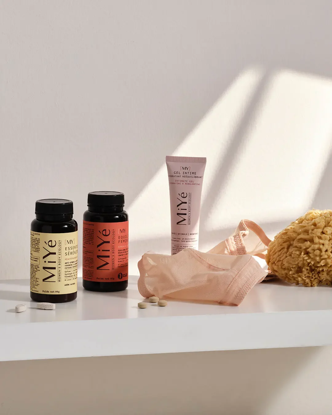 Produits de soins féminins, y compris des compléments alimentaires, un gel intime, un tissu en maille, un distributeur d'eau naturel, des comprimés, et une éponge de bain sur une surface blanche, avec une lumière naturelle créant une ombre douce.