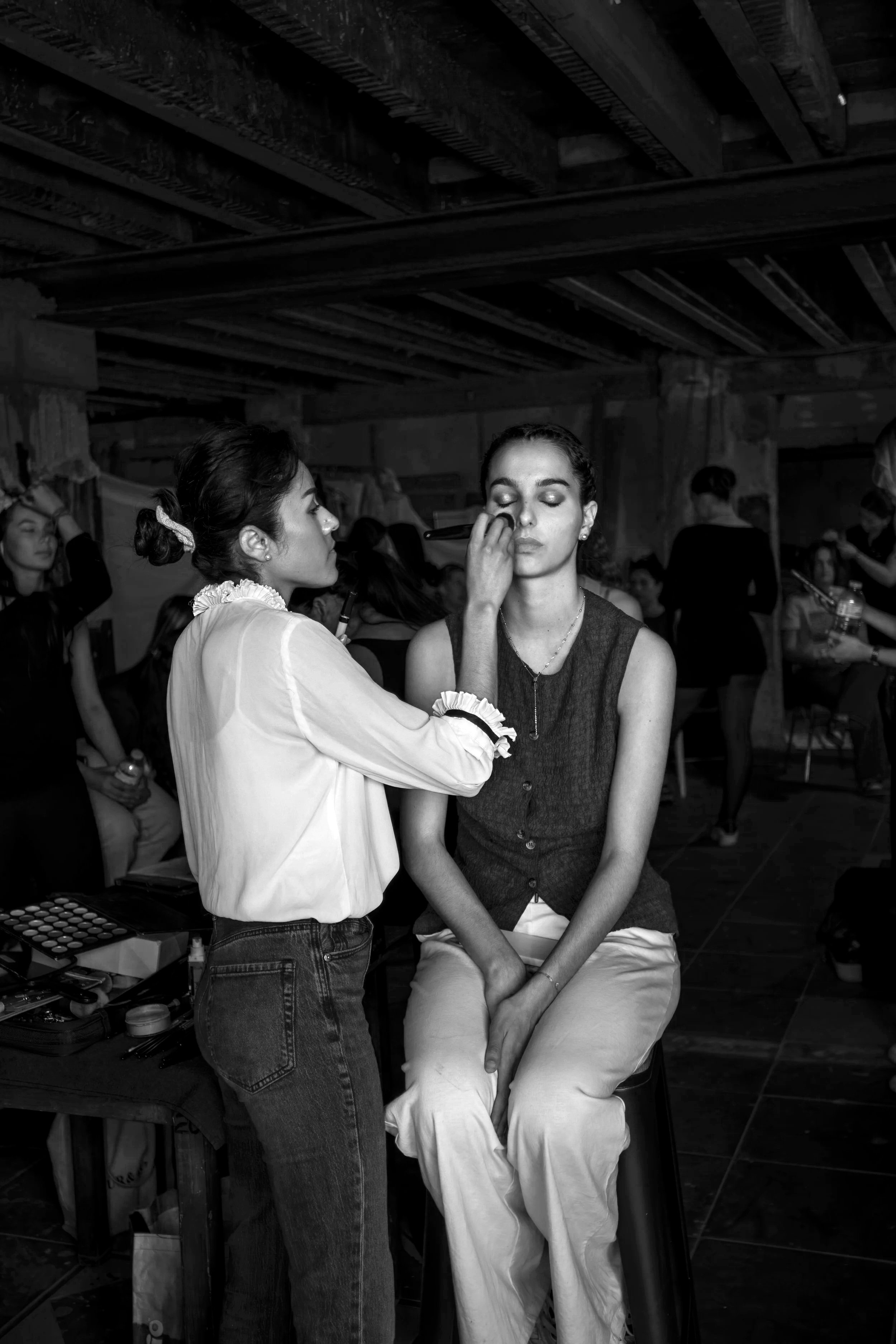 Une femme autre femme, semble-t-il, appliquant du maquillage sur le visage de la femme assise. D'autres personnes sont en arrière-plan, dans un espace intérieur, probablement en train de se préparer ou de faire un shooting photo.