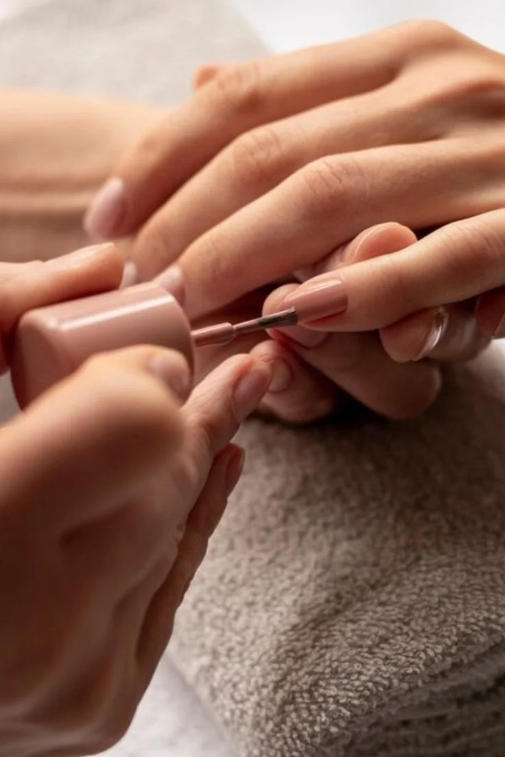 Une personne fait une manucure, en appliquant du vernis à ongles d'une couleur neutre sur ses ongles. La scène montre l'application précise du vernis.