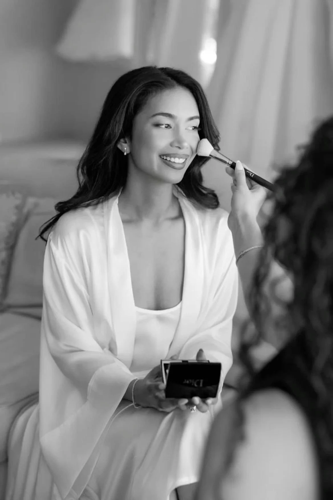 Une femme souriante faisant un maquillage avec un pinceau lors d'une séance de beauté, vue en noir et blanc.