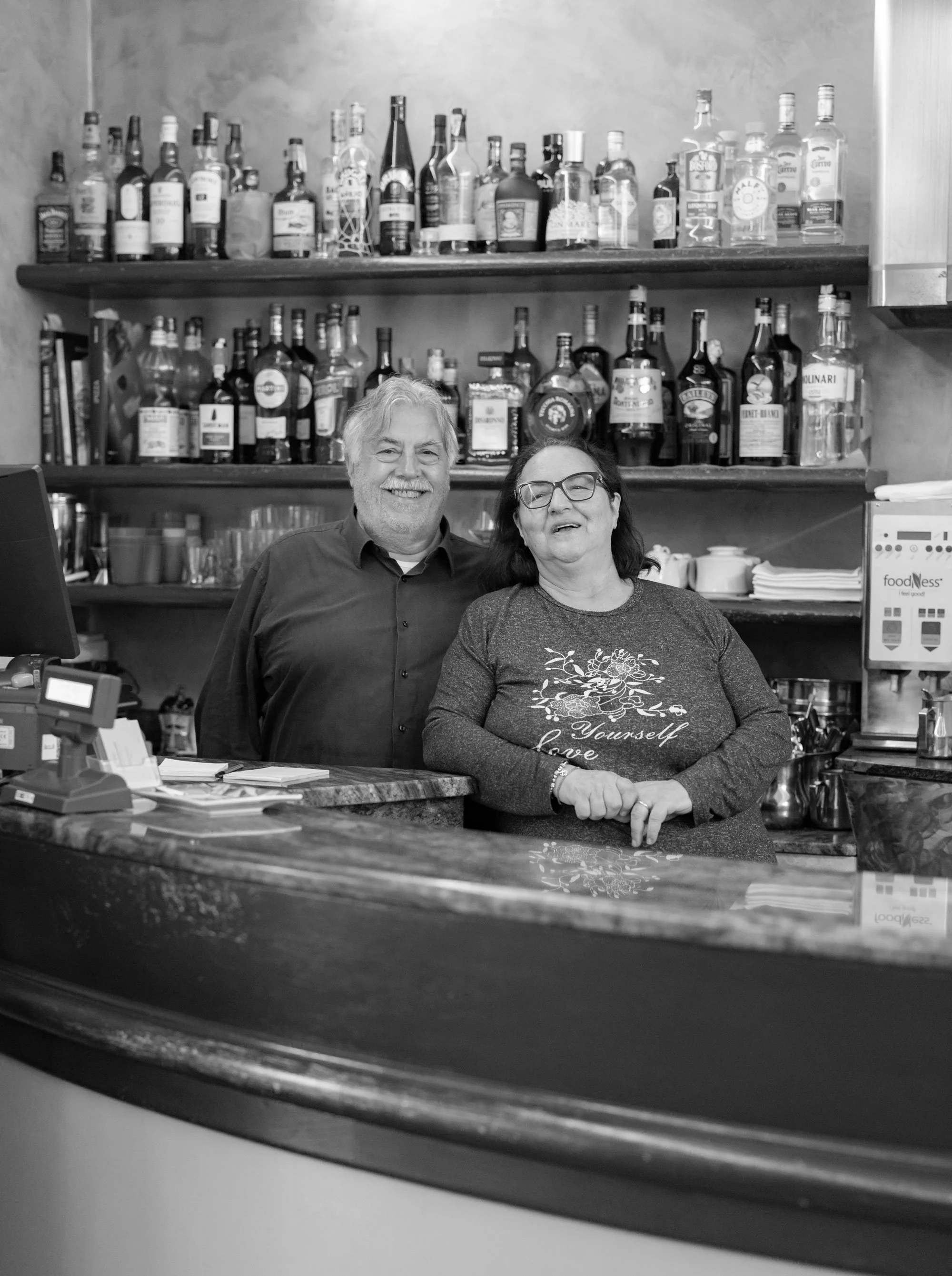 Due persone sorridenti dietro il bancone di un bar o ristorante, con scaffali pieni di bottiglie di alcolici sullo sfondo, in un ambiente interno.