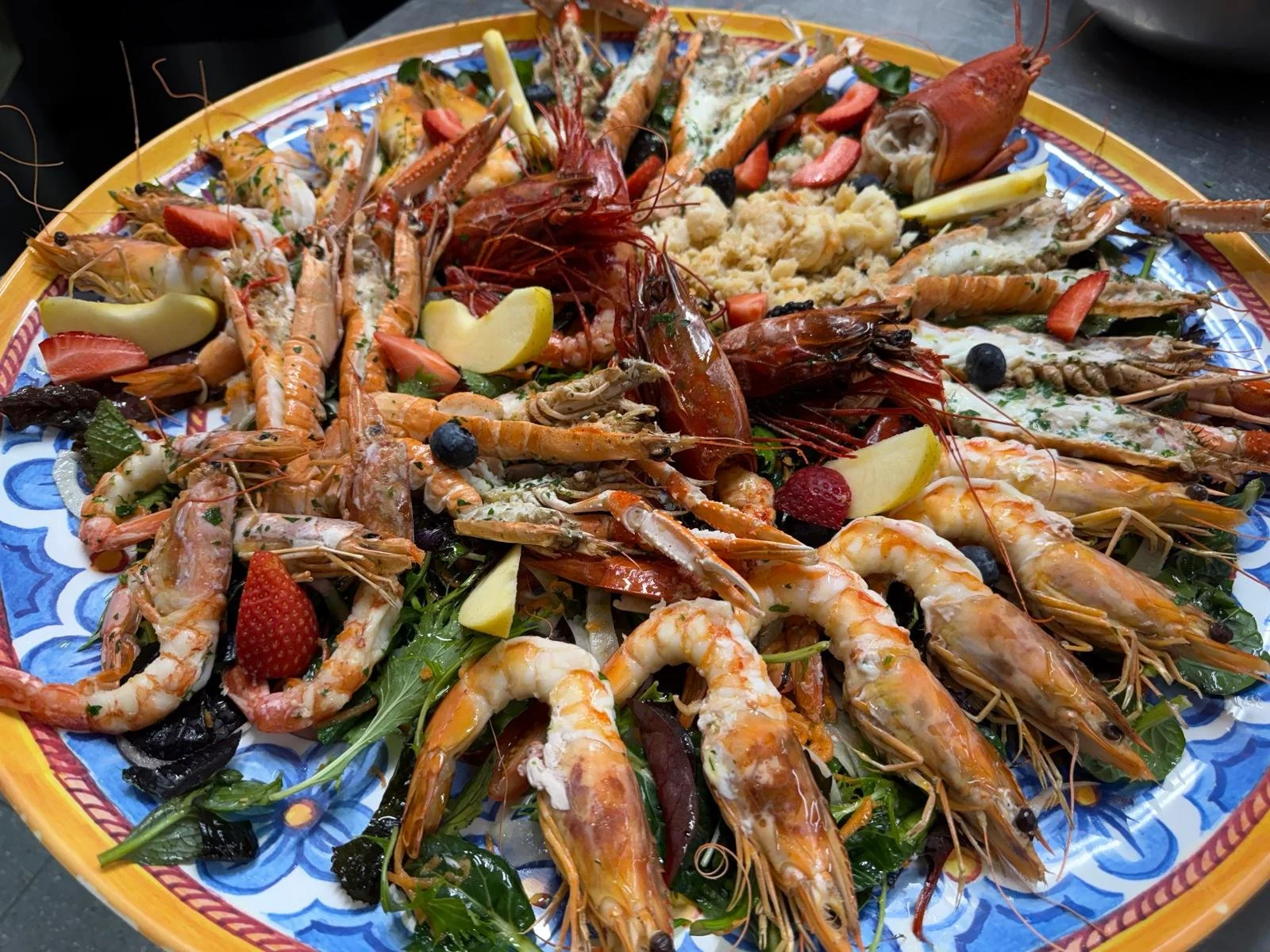 Piatti di frutti di mare freschi, tra cui gamberi, aragoste, e altri crostacei, decorati con frutta, foglie verdi e verdure.