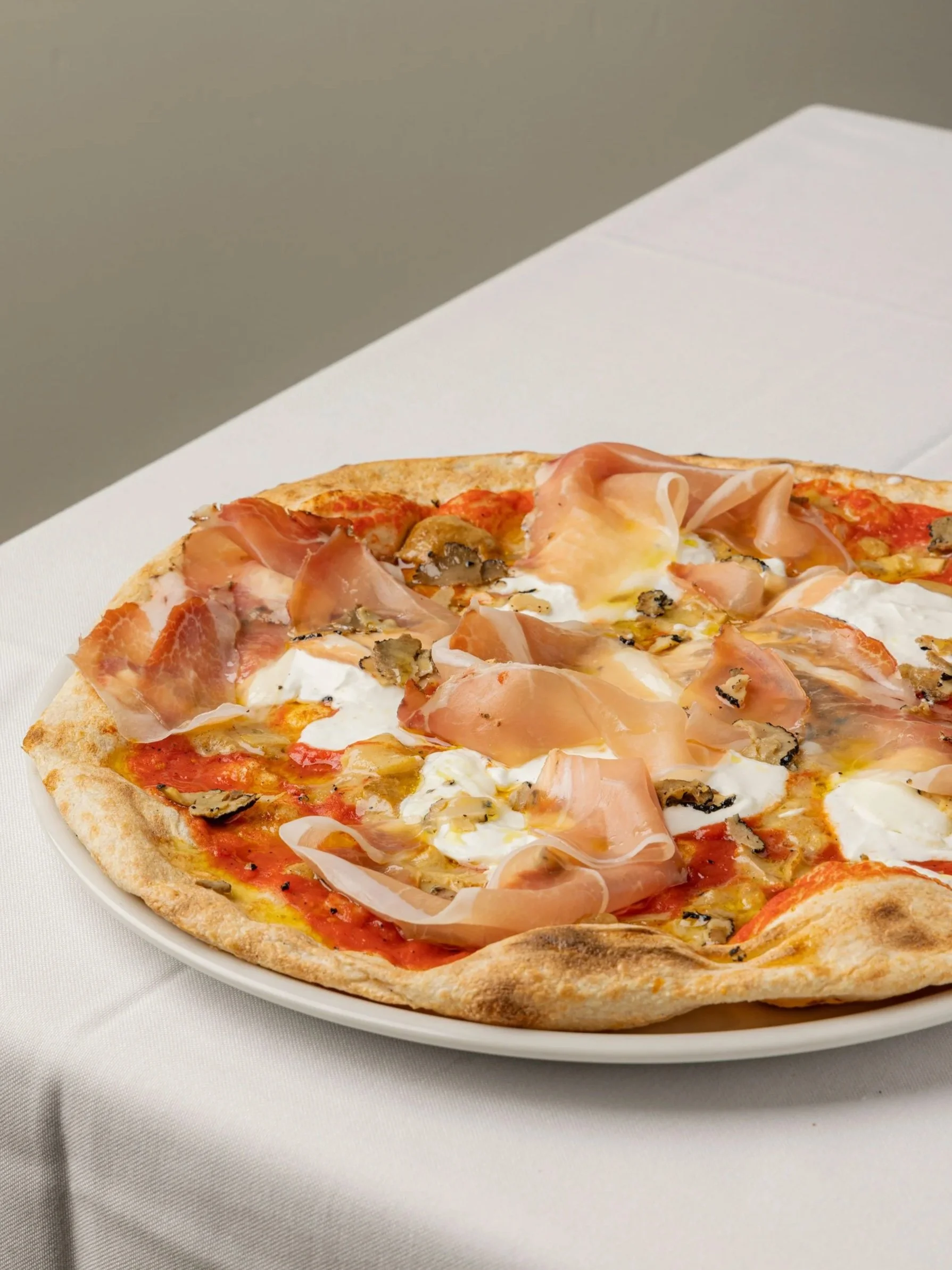 Una pizza con prosciutto, funghi e mozzarella su piatto bianco su tovaglia di tessuto bianca.