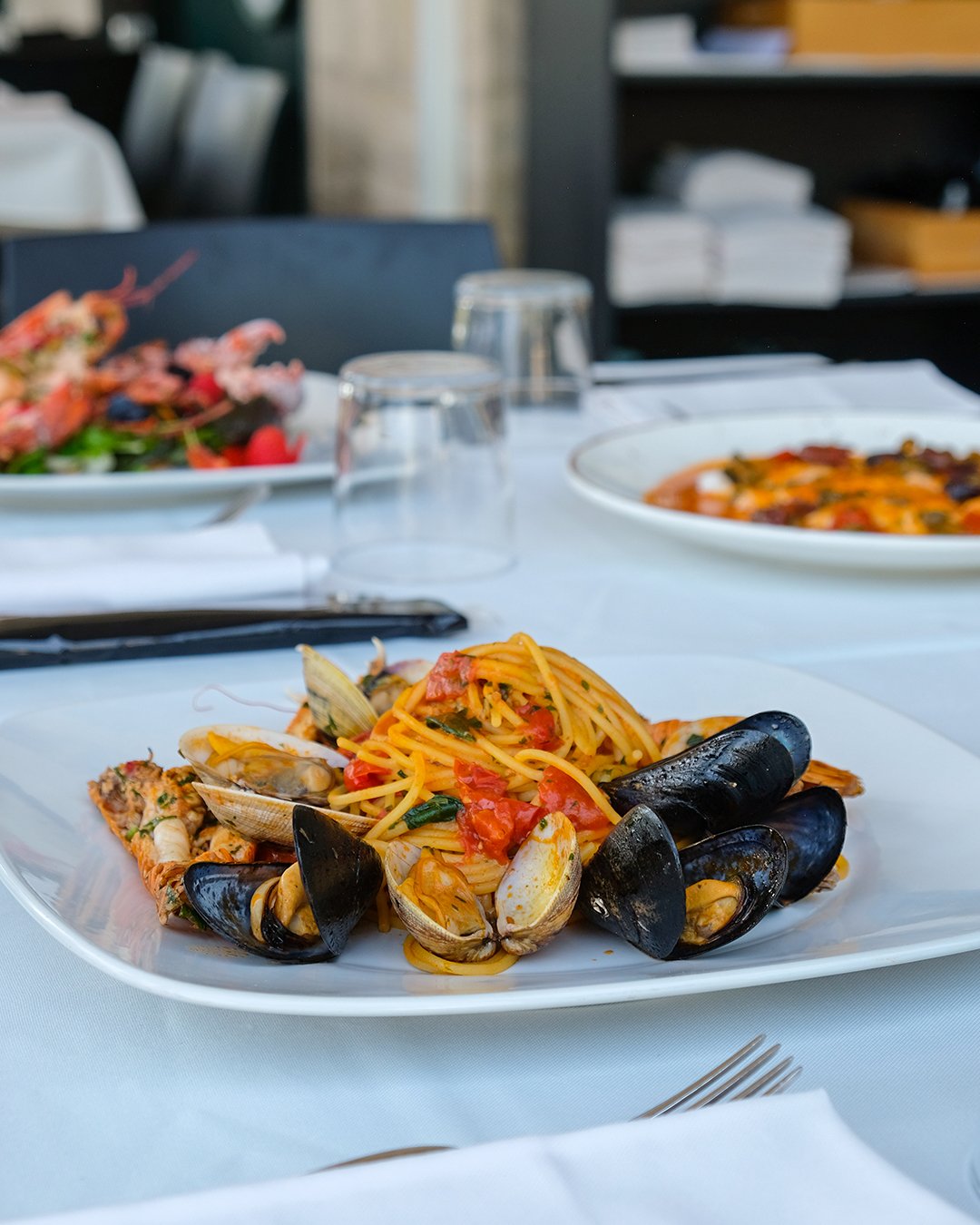 Piatti di spaghetti alle vongole con pomodoro e basilico, serviti su un piatto bianco su una tovaglia bianca, con bicchieri di acqua e altri piatti sul tavolo.