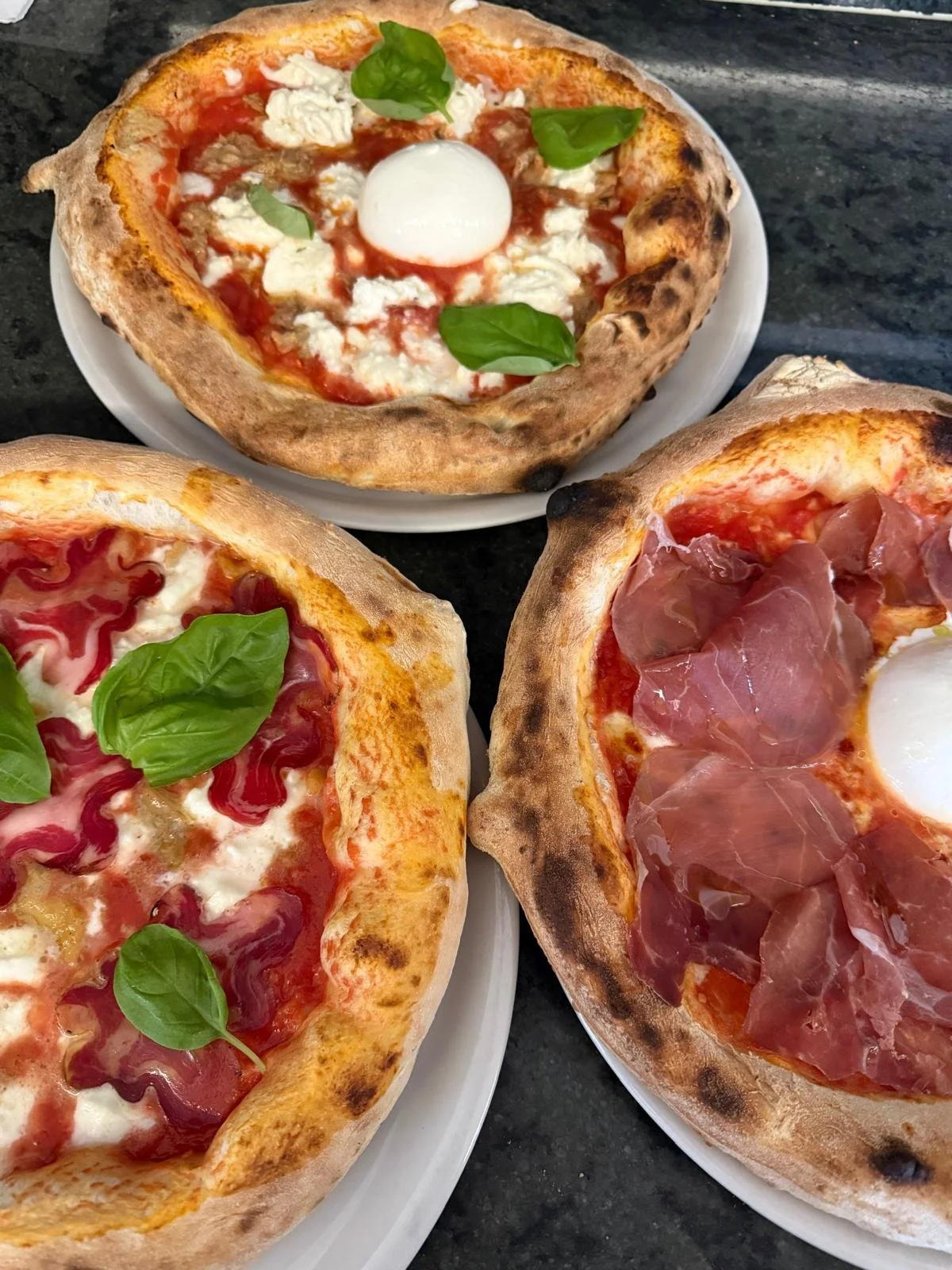 Tre pizze con vari condimenti come prosciutto, mozzarella, pomodoro e basilico, in teglie di pietra, su un tavolo di marmo.