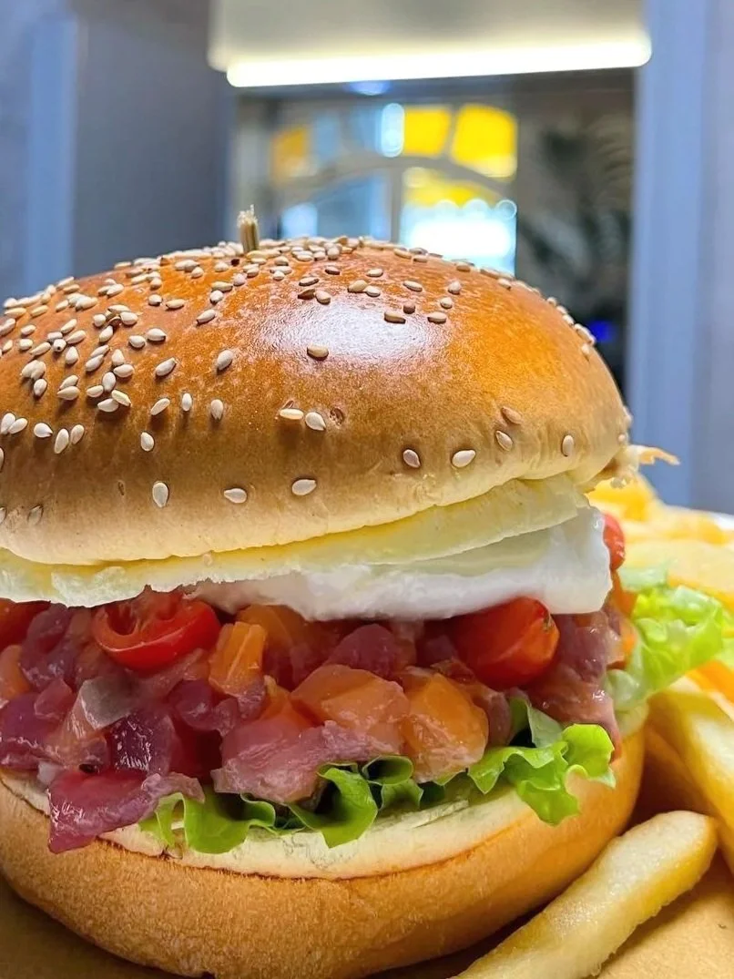 Un panino con hamburger con insalata, pomodori, un uovo fritto e un panino con semi di sesamo, accompagnato da patatine fritte.