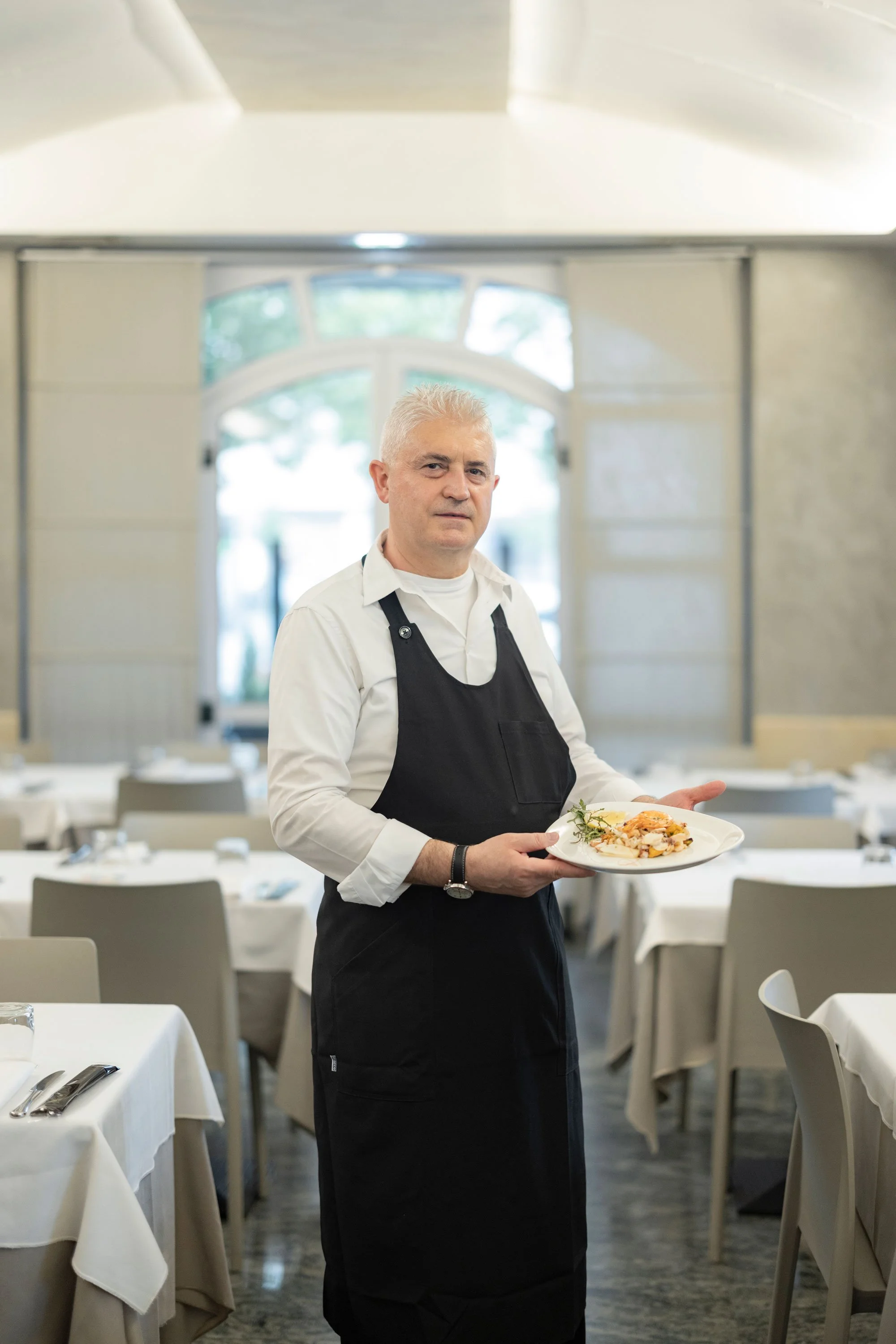 Uomo con capelli grigi e camicia bianca, indossando grembiule nero, tiene un piatto con cibo in un ristorante elegante arredato con tavoli ricoperti da tovaglie bianche.