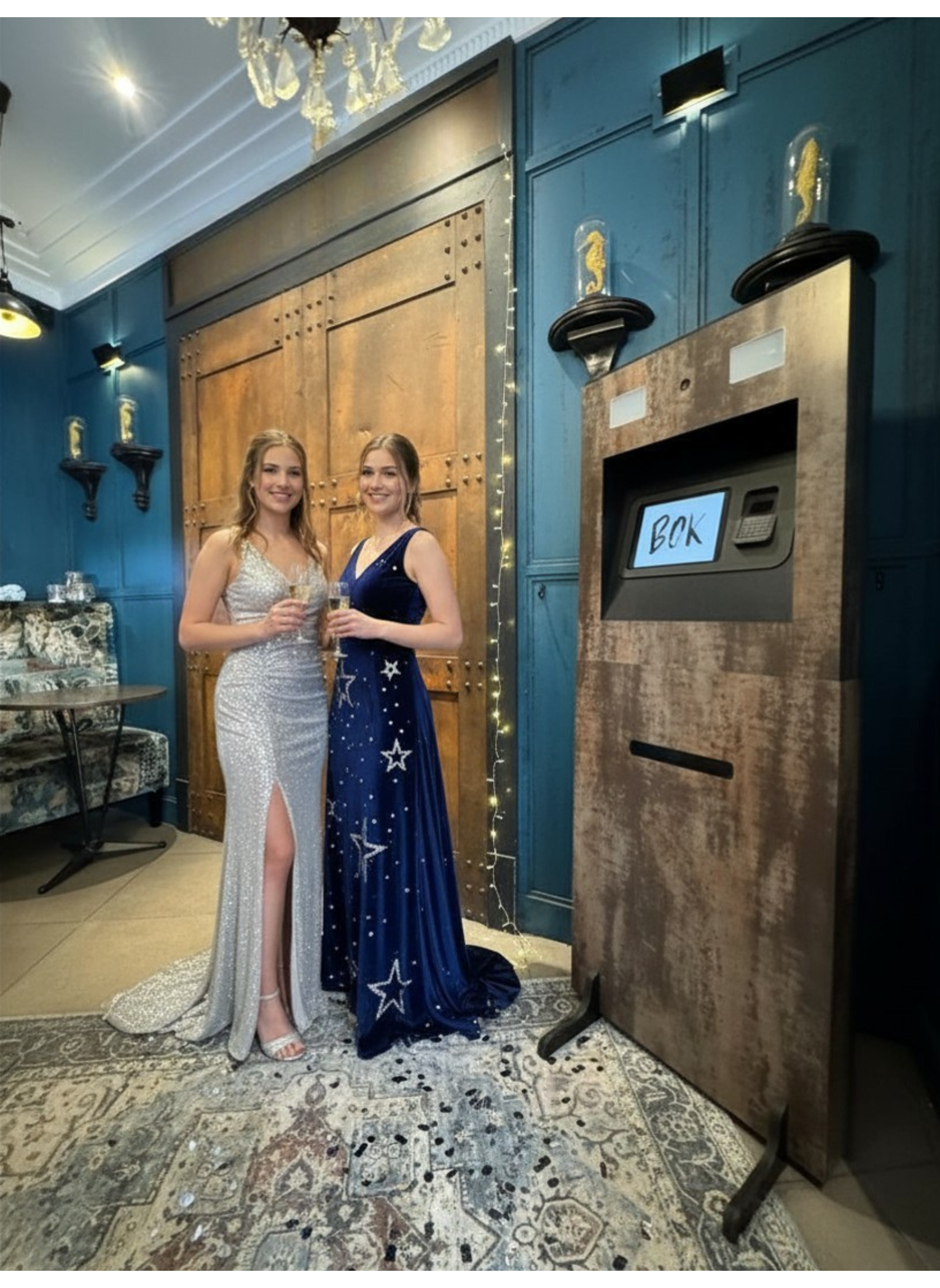 Deux femmes habillées en robes de soirée élégantes, tenant des verres, pourraient être à une fête ou un événement formel dans un intérieur décoré avec des meubles modernes et des murs bleus.