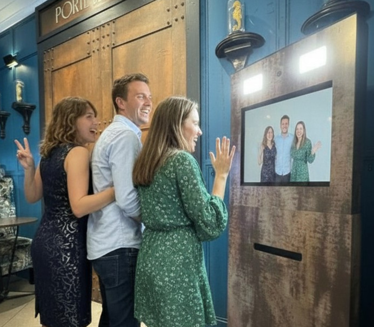 Quatre personnes souriantes sont regroupées devant un écran vidéo dans un restaurant, posent pour une photo virtual 'selfie'.