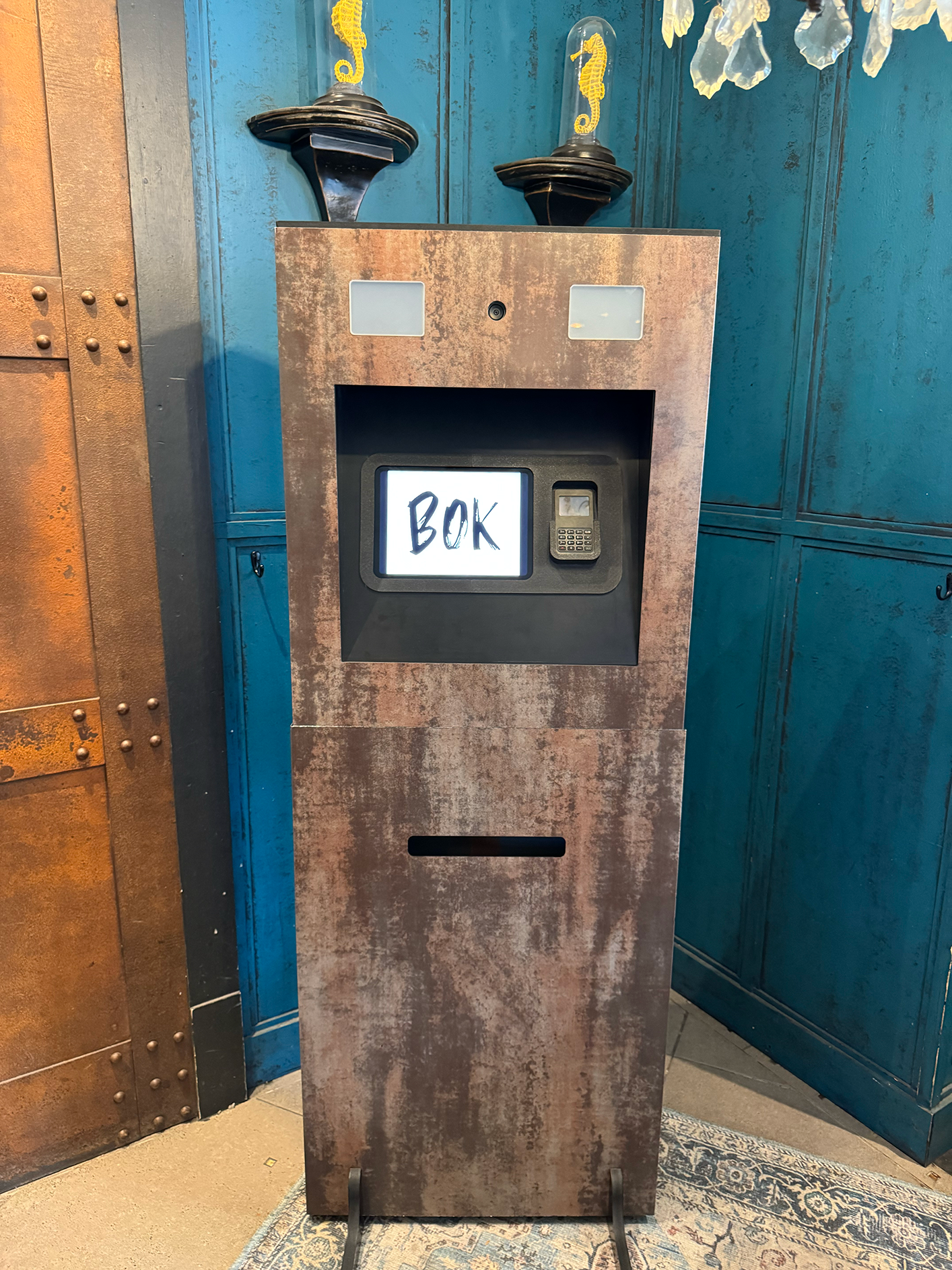 Un distributeur automatique avec l'inscription «BOK» affichée sur un écran, situé dans une pièce avec des murs en bleu et en cuivre. Le distributeur possède un clavier et un écran tactile.
