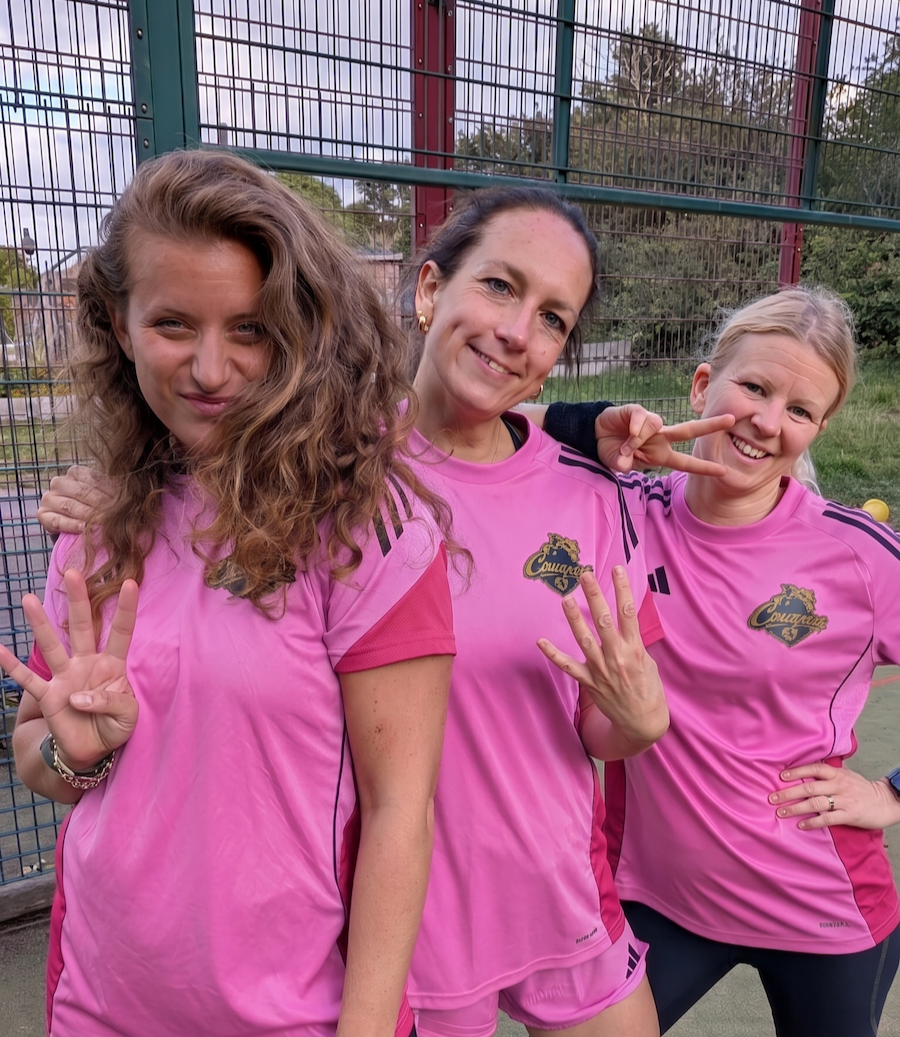 Three women in pink sports uniforms posing outdoors, making a 4-4-2 gesture with their hands, in front of a tall wire fence and green trees.