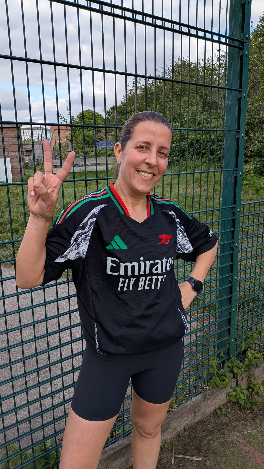 A woman in a black sports jersey making a peace sign with her right hand and smiling, outdoors next to a green metal fence.
