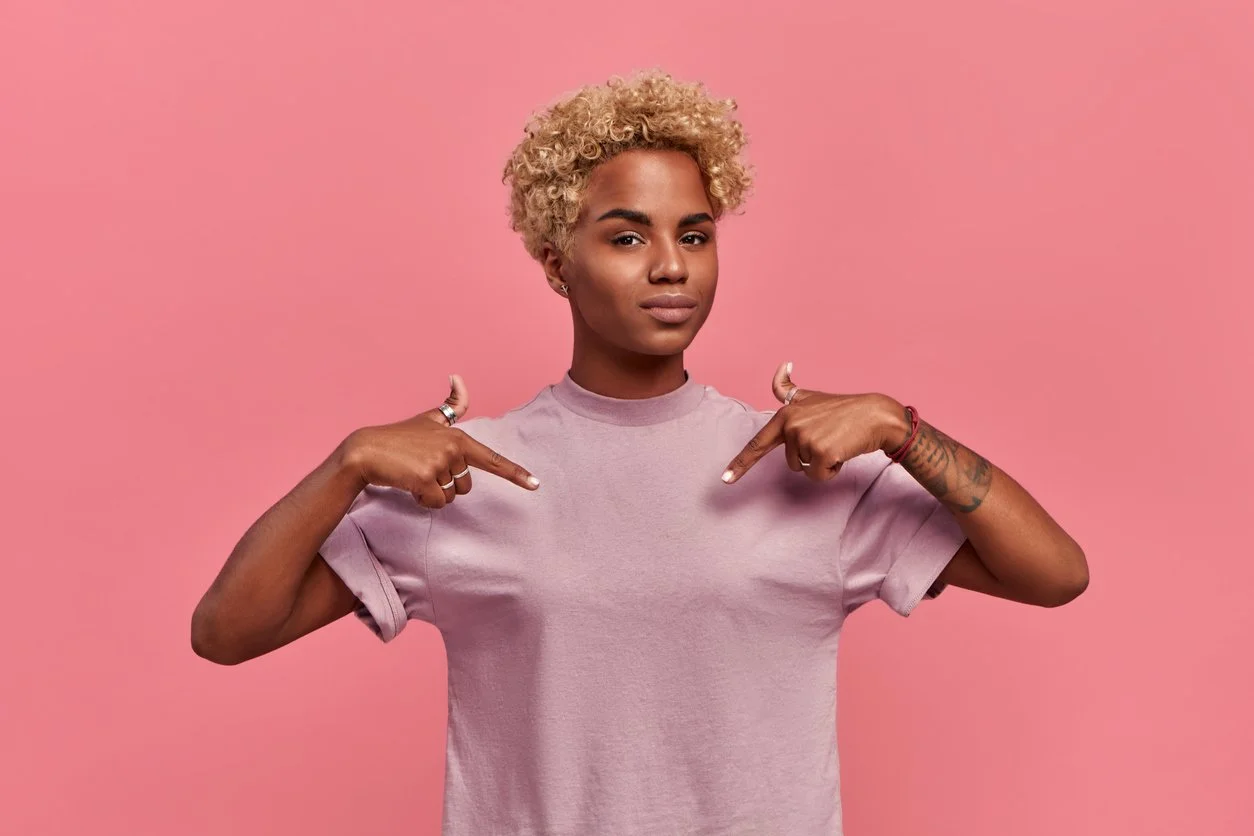 A woman with short curly blonde hair pointing to their chest against a pink background.