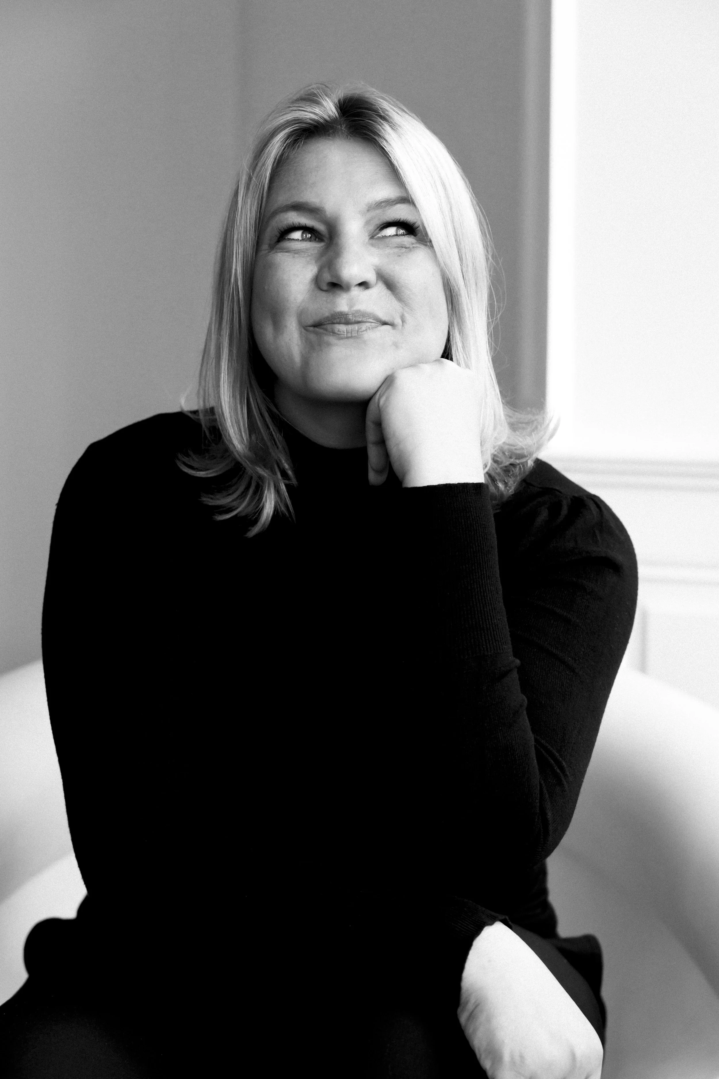 Schwarz-Weiß-Foto einer lächelnden Frau mit blonden Haaren, die nach oben schaut, die rechte Hand unter dem chin auf dem Gesicht ruhend, sitzend vor einer hellen Wand.