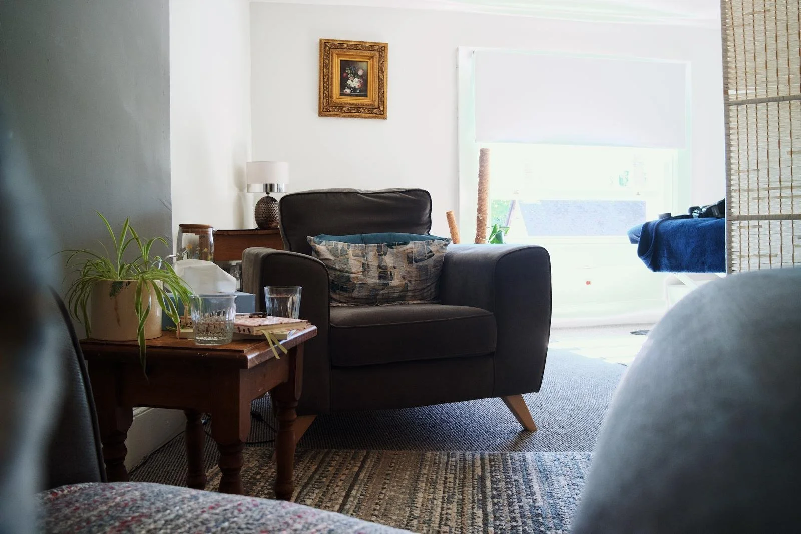 Living room with a black armchair, a wooden side table with a plant and glasses, a lamp, a wall painting, a window with a white blind, and various objects including a blue towel and a camera.