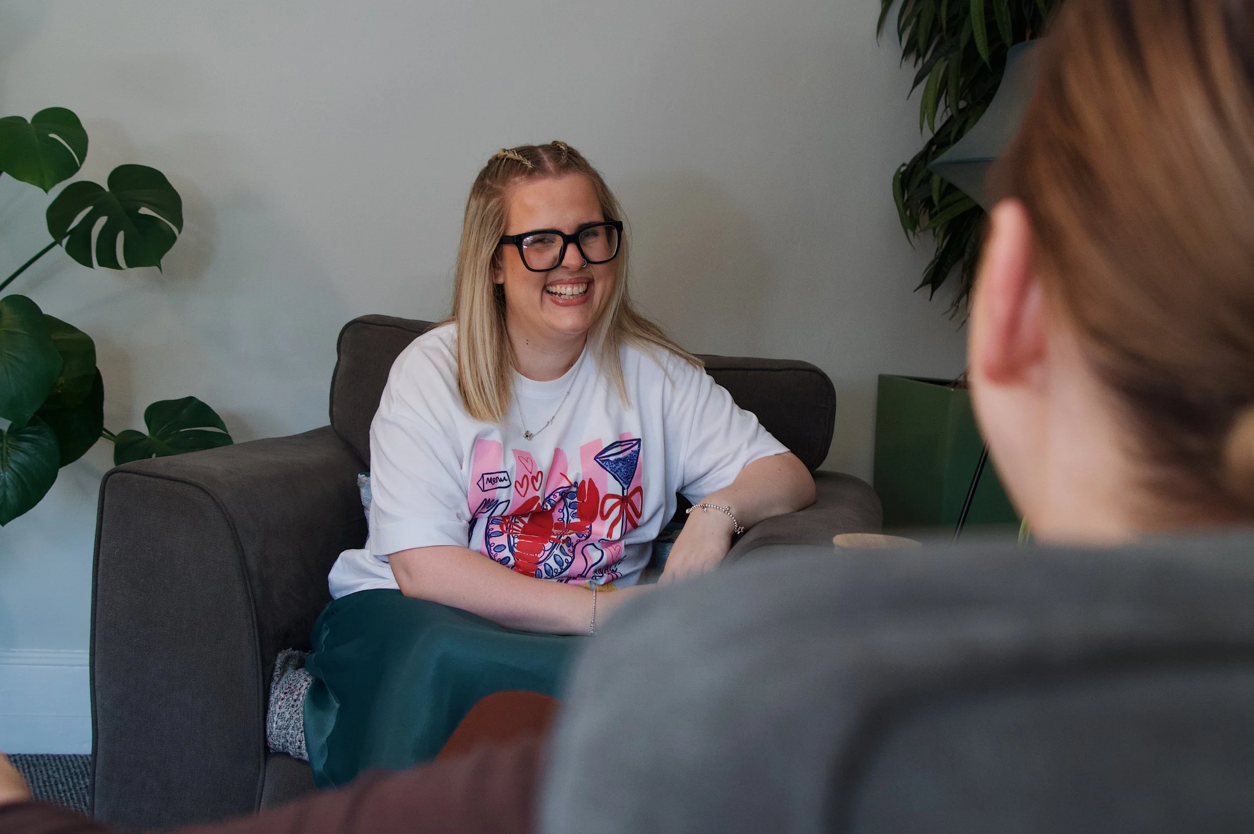 A woman with blonde hair, glasses, and a white T-shirt smiling and talking to a person with red hair, in a room with large green plants.