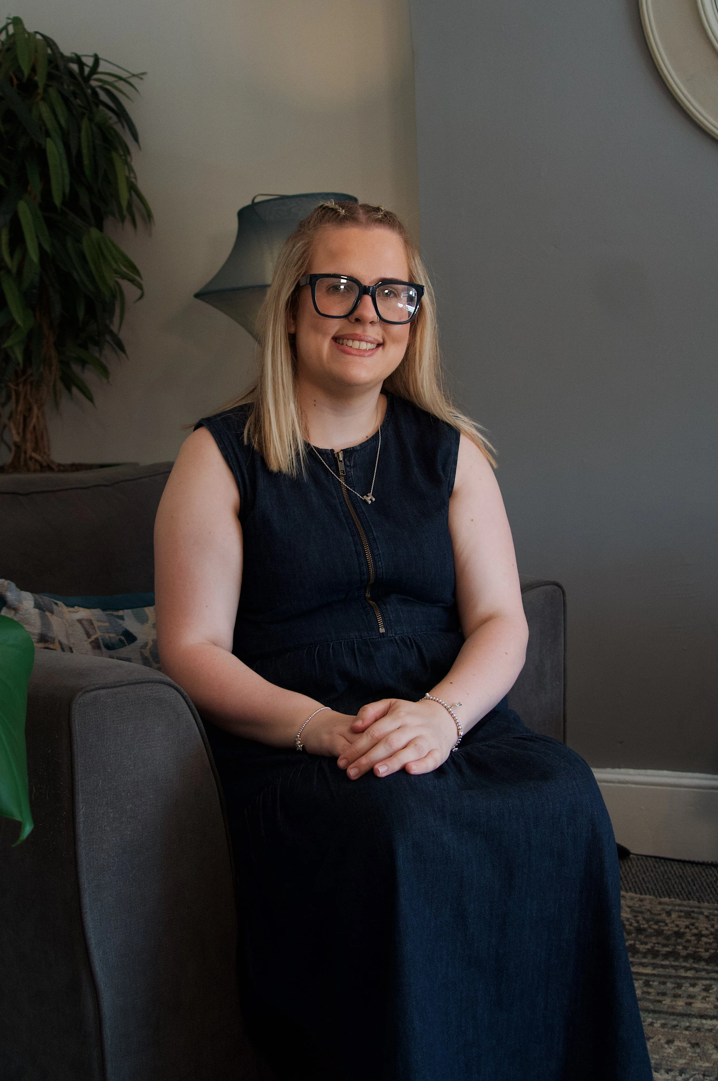 A woman with blonde hair wearing glasses, a black sleeveless dress, and jewelry, sitting on a gray couch with a plant and lamp in the background.