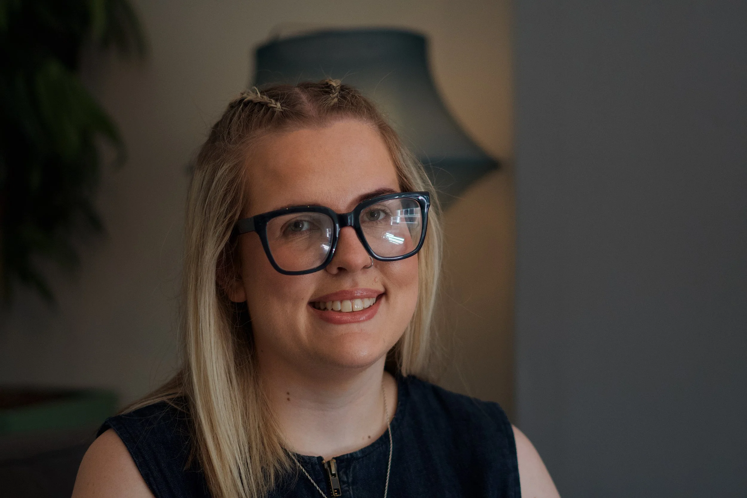 A young woman with blonde hair, glasses, and a black sleeveless top smiles indoors, with a blurred background and a lamp.