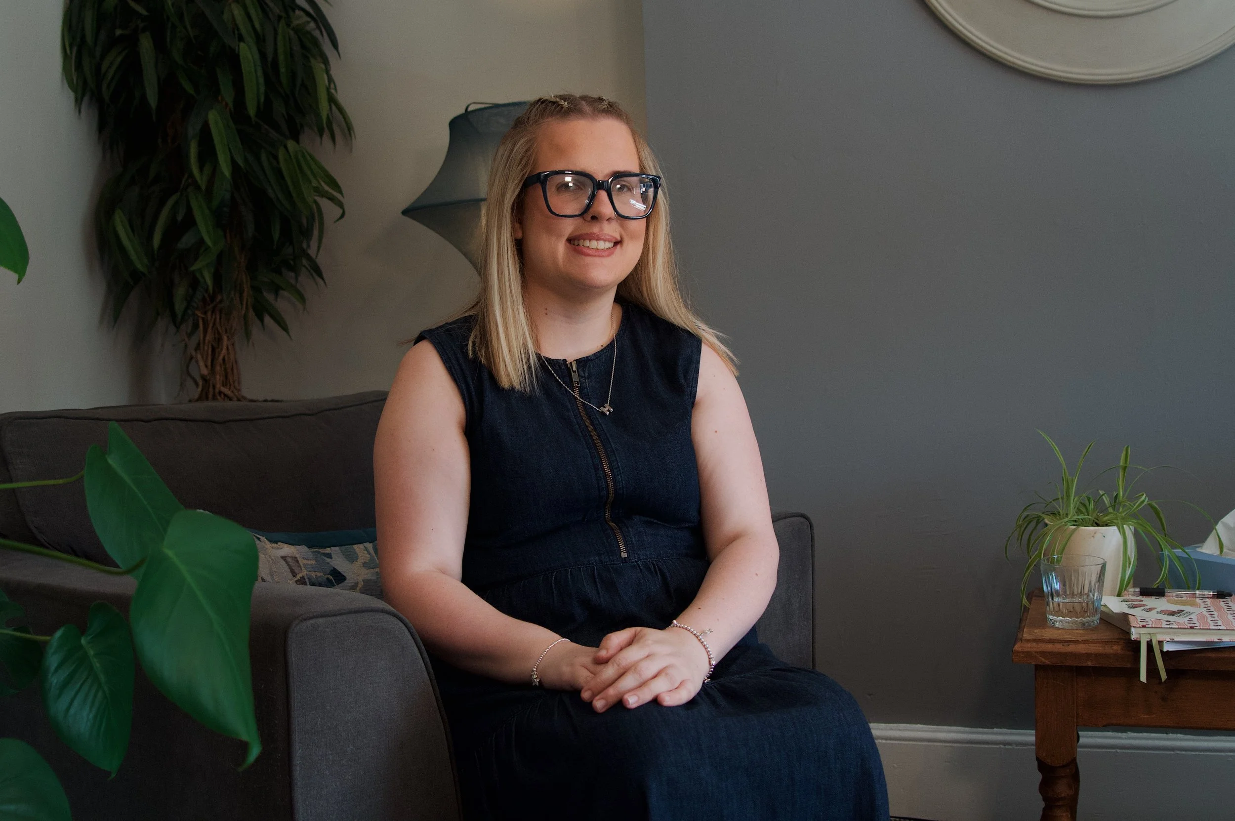 A woman with blonde hair, glasses, wearing a sleeveless black dress, seated on a gray couch in a living room with a potted plant to her left, a table with a plant, glasses of water, and magazines to her right, and a lamp behind her.