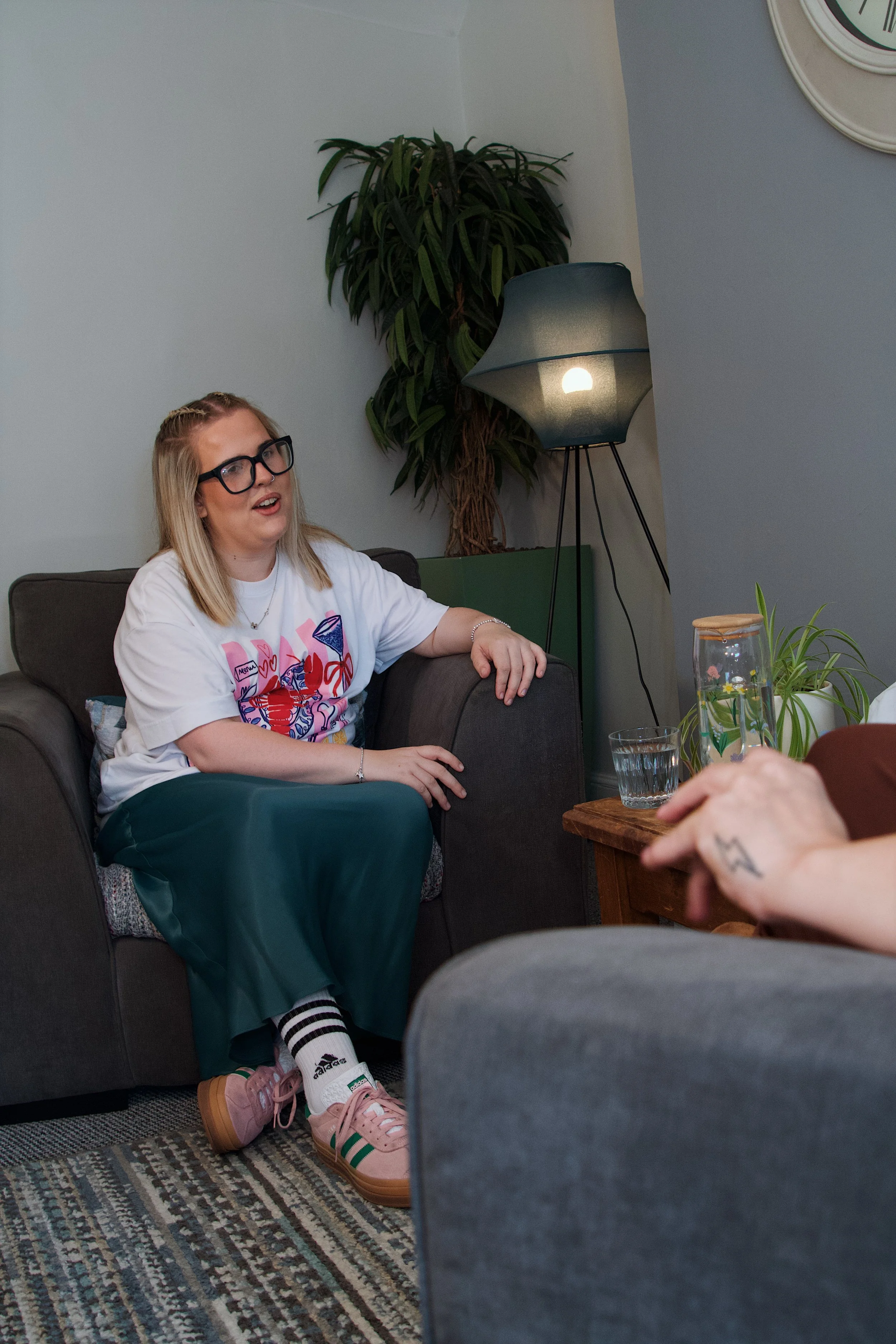 A woman with blonde hair, glasses, and a white t-shirt with colorful graphics is sitting on a dark grey couch, smiling and talking. She has one arm resting on the armrest. Next to her is a wooden table with a glass of water, a glass jar with a lid, and some greenery inside. Behind her, there is a tall green plant and a modern floor lamp with a dark shade. The room has light-colored walls and a patterned rug on the floor.