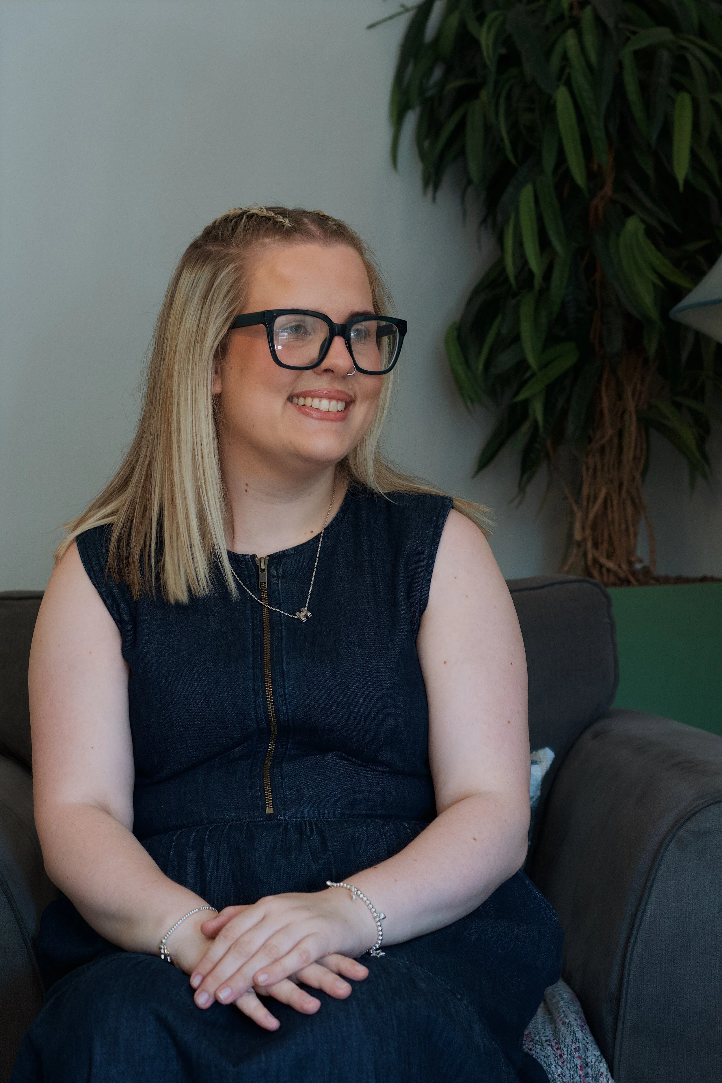 A smiling young woman with blonde hair, wearing black glasses, a nose ring, a silver necklace, and bracelets on her wrists, sitting on a dark couch in a room with light-colored walls and a large green plant in the background.