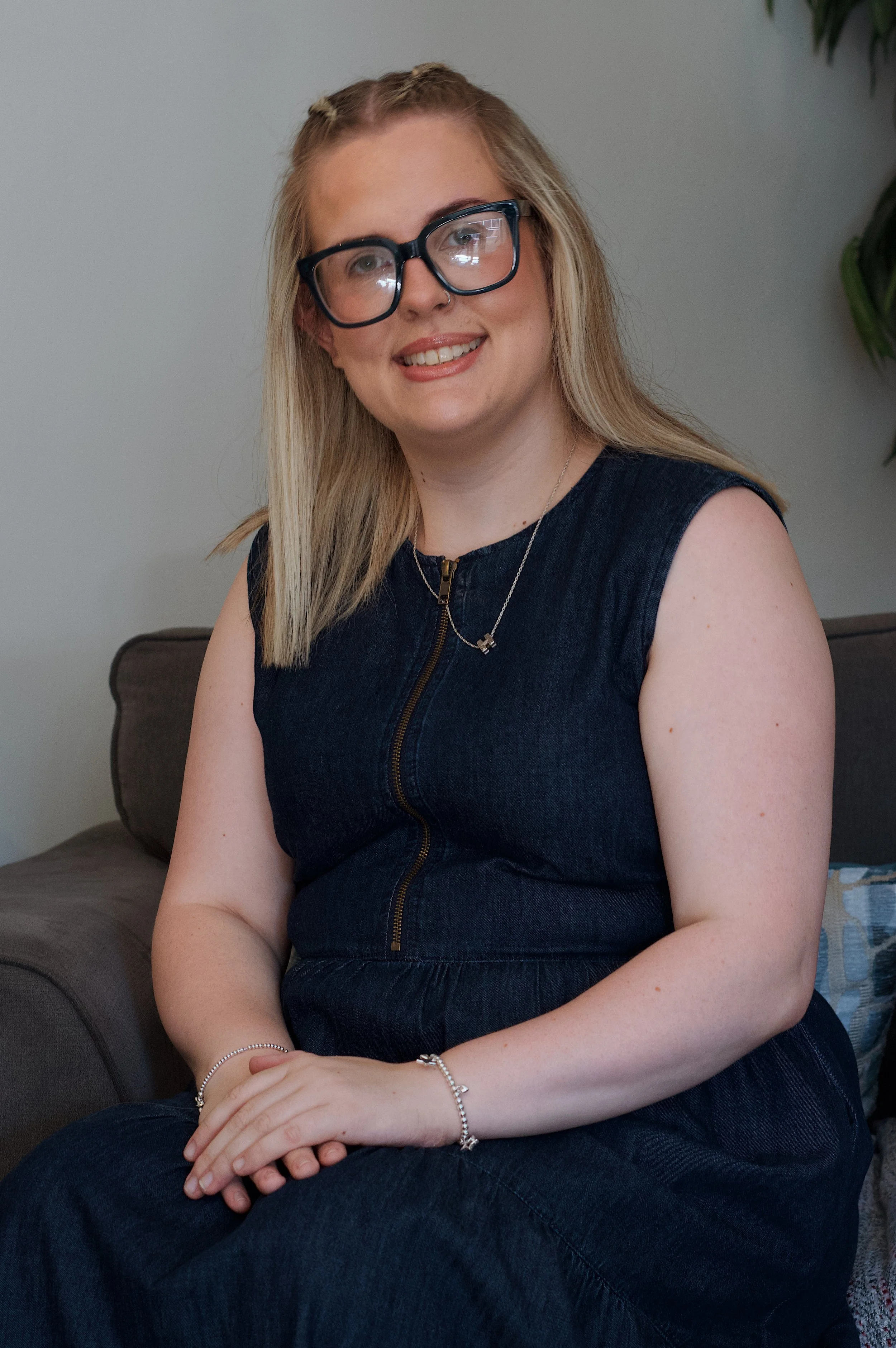 A young woman with blonde hair, wearing large black glasses, a black sleeveless zip-up dress, and jewelry including rings, a necklace, and bracelets, sitting on a couch in a room with a gray wall and a leaf of a plant visible in the background.