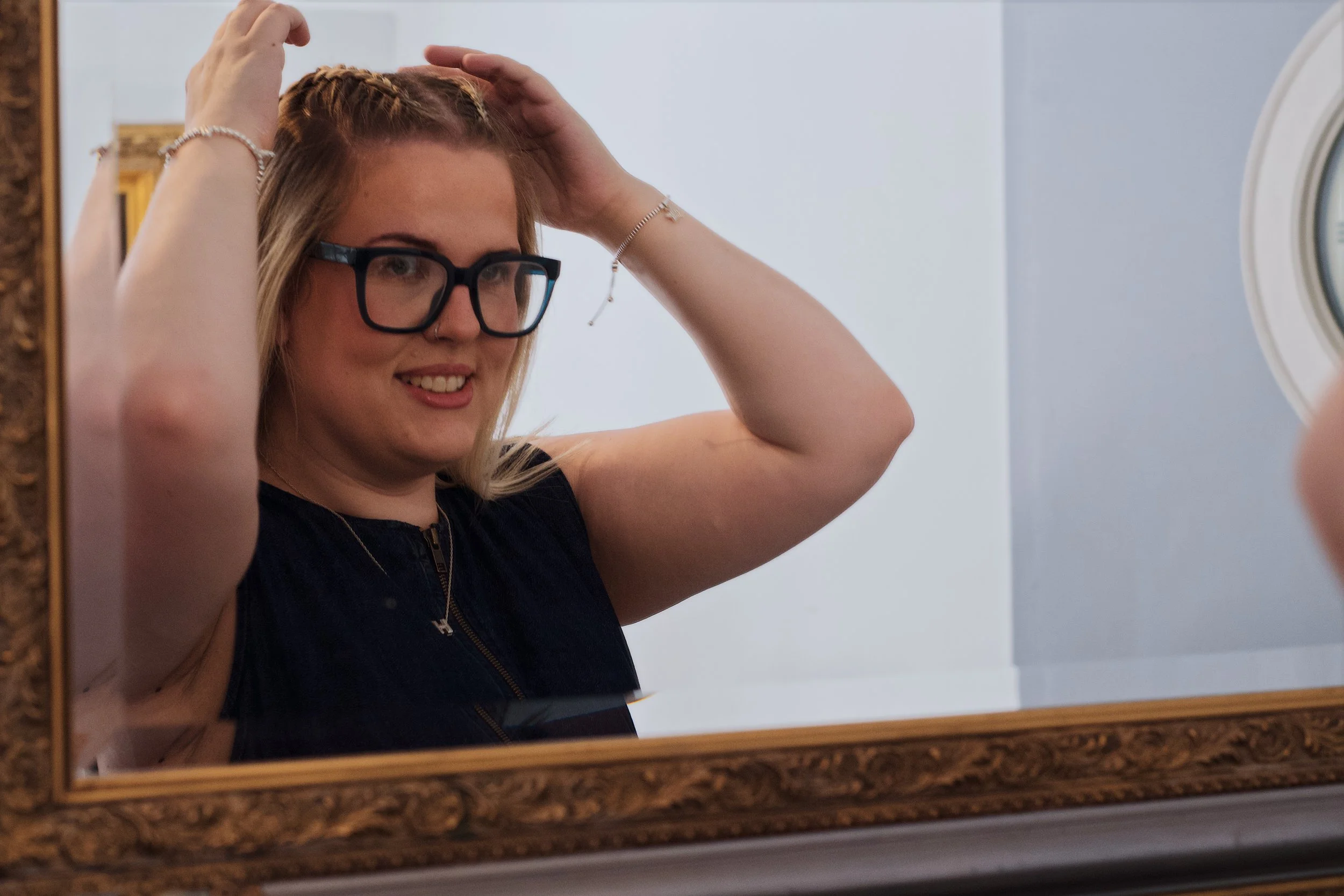 A woman with glasses smiling at a mirror, adjusting her hair with both hands.