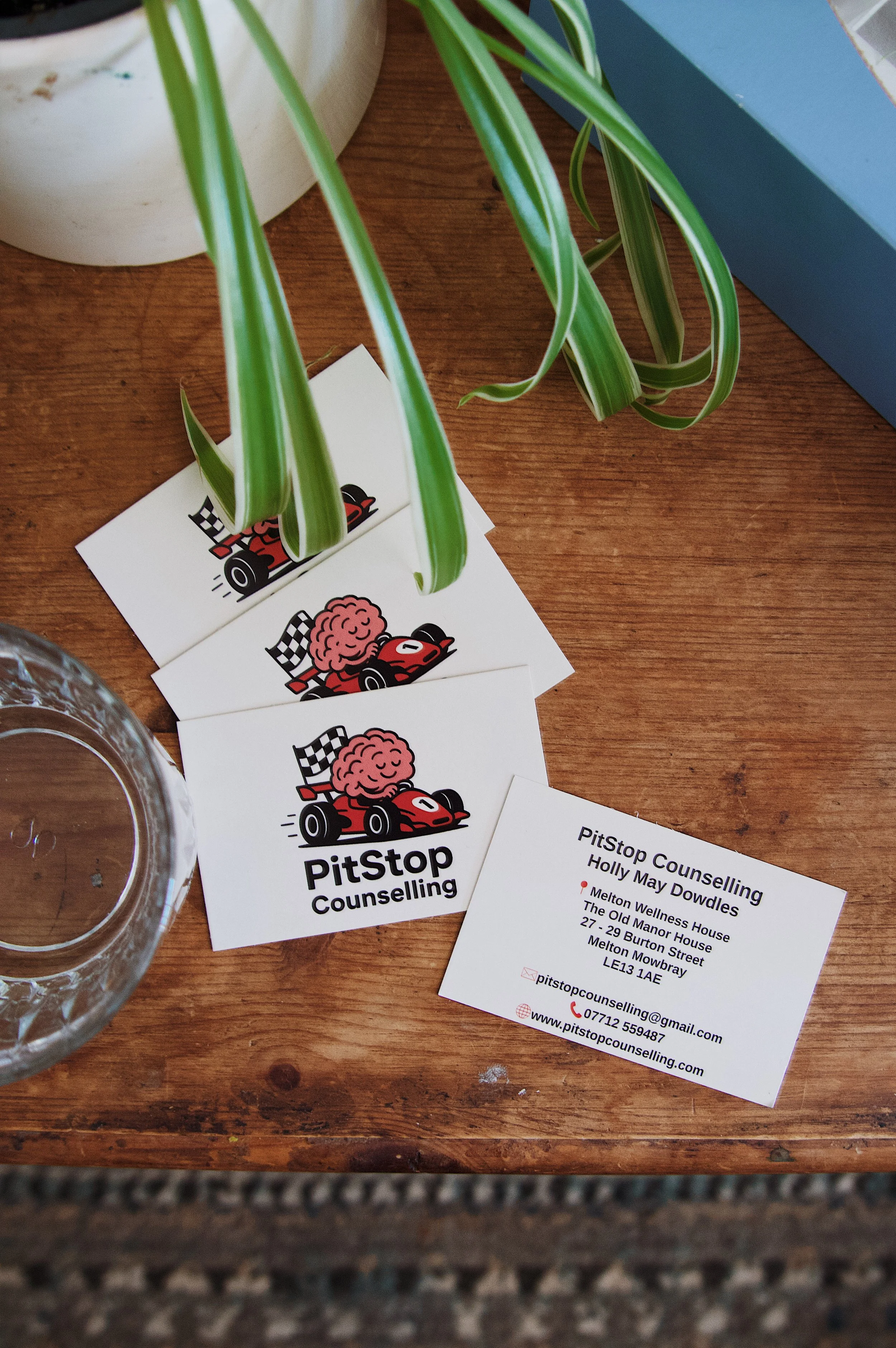 Business cards for PitStop Counselling with a racing car and brain logo, placed on a wooden table next to a potted plant and a glass of water.