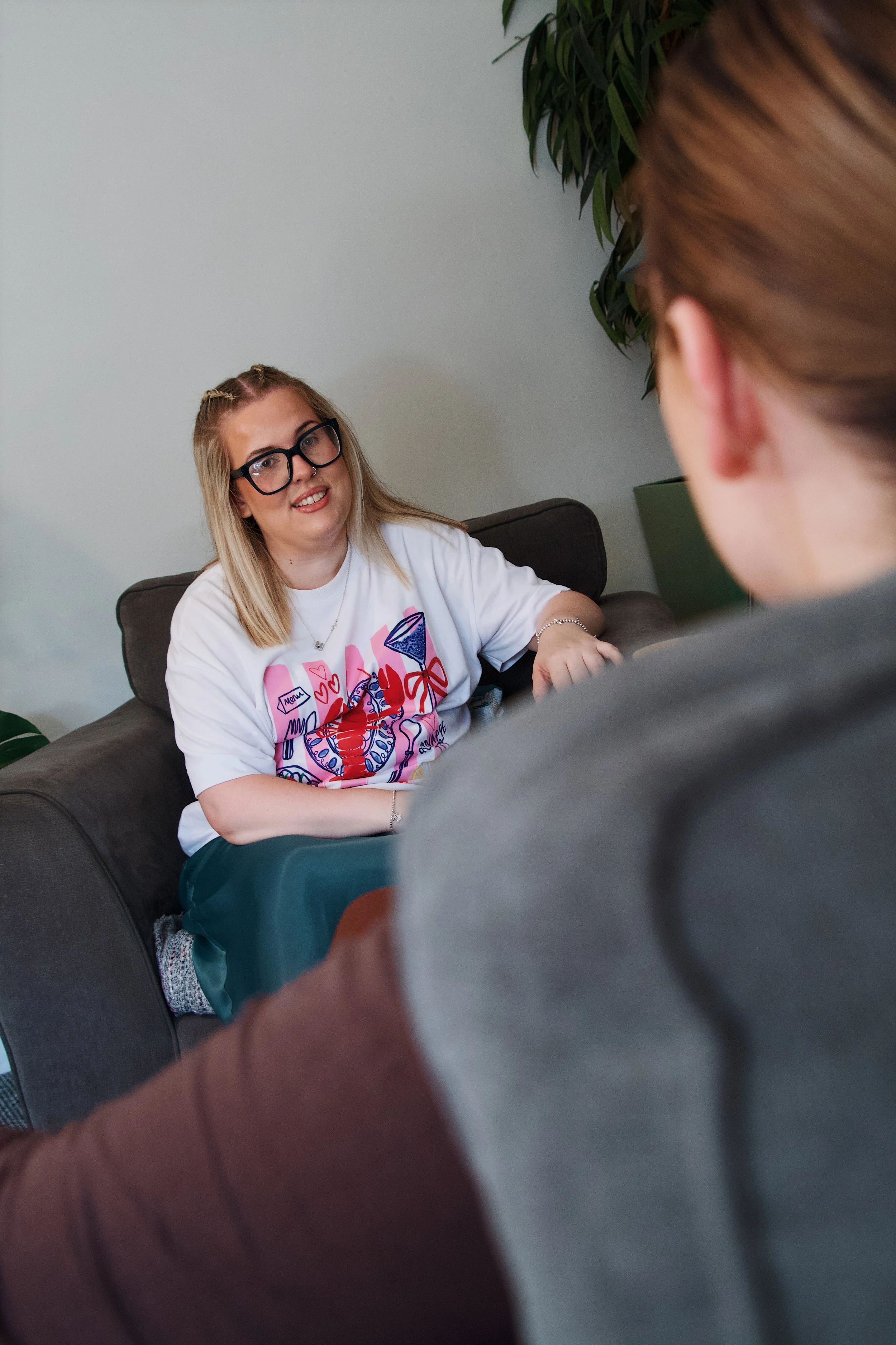 Two women sitting on a couch, engaged in conversation. The woman on the left is wearing glasses and a colorful t-shirt, with blonde hair and a smile. The woman on the right has her back to the camera, with red hair pulled back. There are plants in the background.