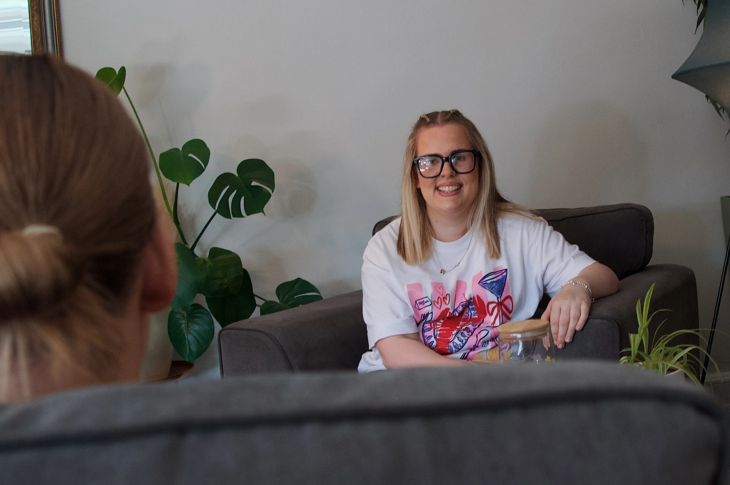 A woman with long blonde hair, glasses, and a white colorful T-shirt sitting on a couch smiling. There are green plants and a lamp in the background. Someone's head with red hair is visible in the foreground.