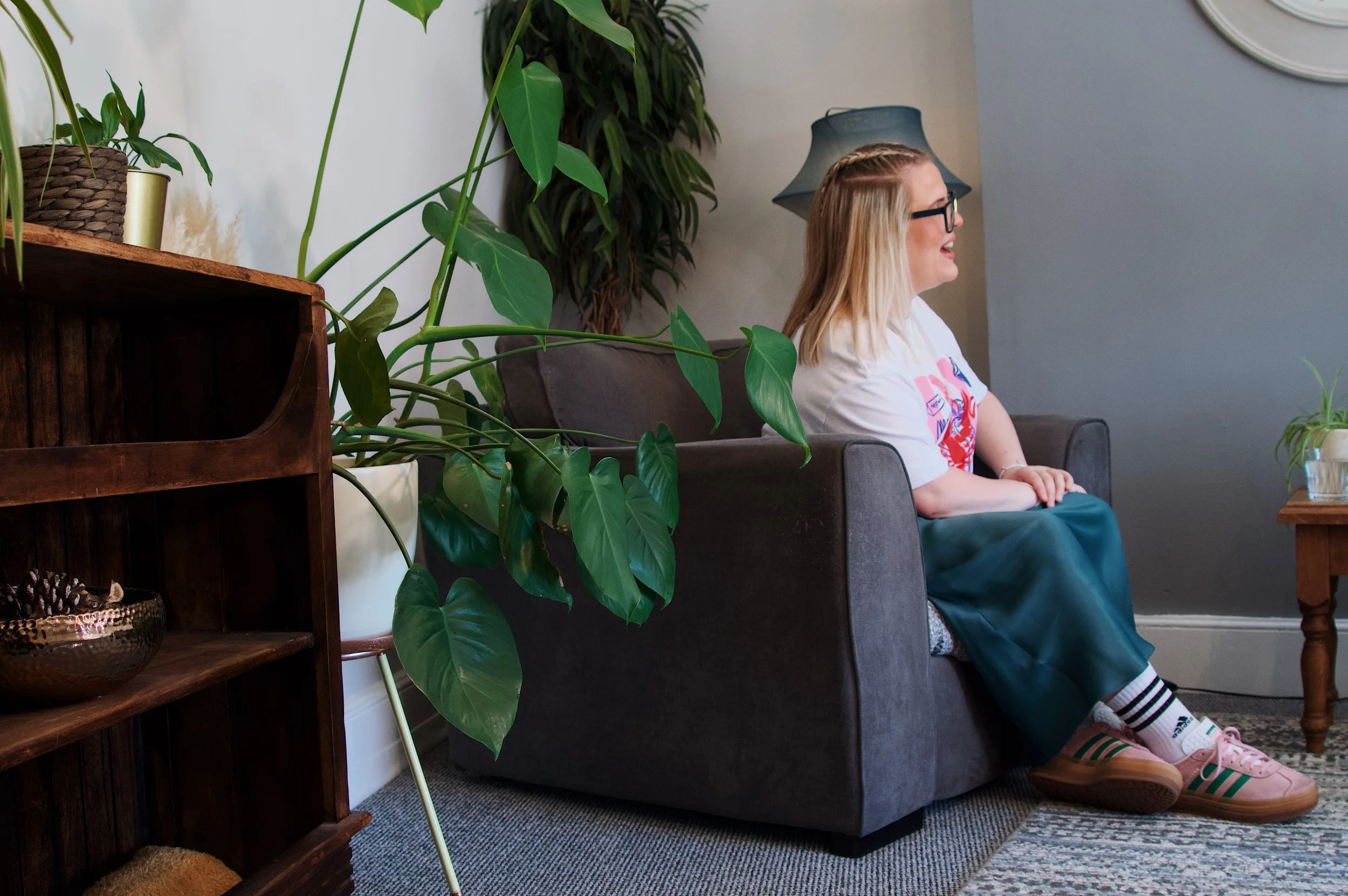 A woman with blonde hair and glasses sitting on a dark gray couch, smiling. She is wearing a white T-shirt with a colorful graphic and teal pants. There are green plants in the foreground and background, and a blue lamp in the background. A wooden shelf with decorative items is on the left side.