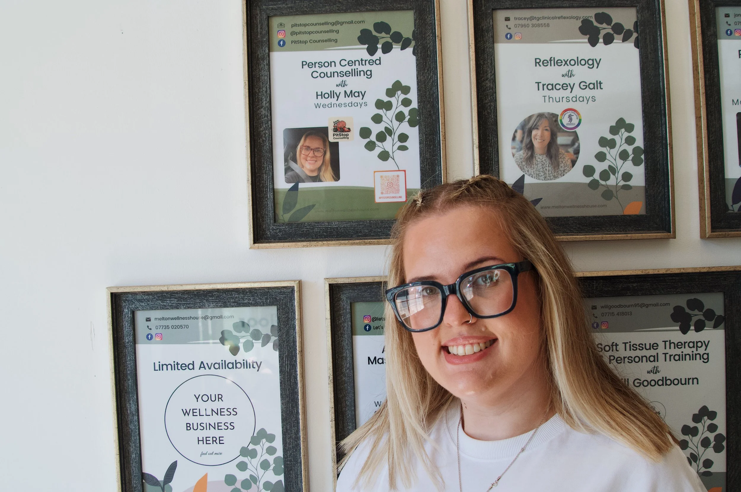 Young woman with blonde hair, glasses, and a nose ring smiling in front of framed posters about wellness and counseling services.