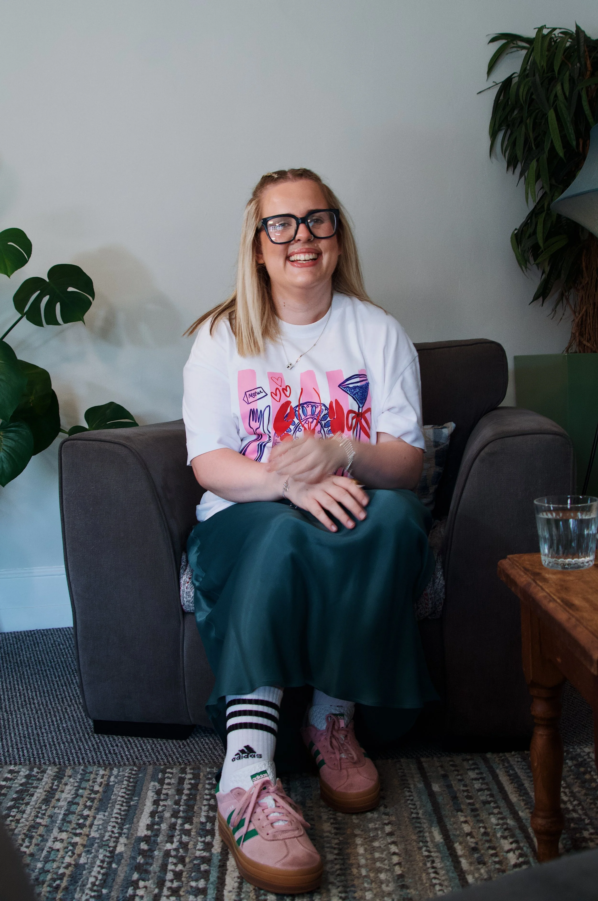 A young woman with blonde hair, glasses, and a nose ring, smiling while seated on a dark gray sofa in a room with green plants. She is wearing a white graphic t-shirt, dark green pants, and pink sneakers, with striped socks.
