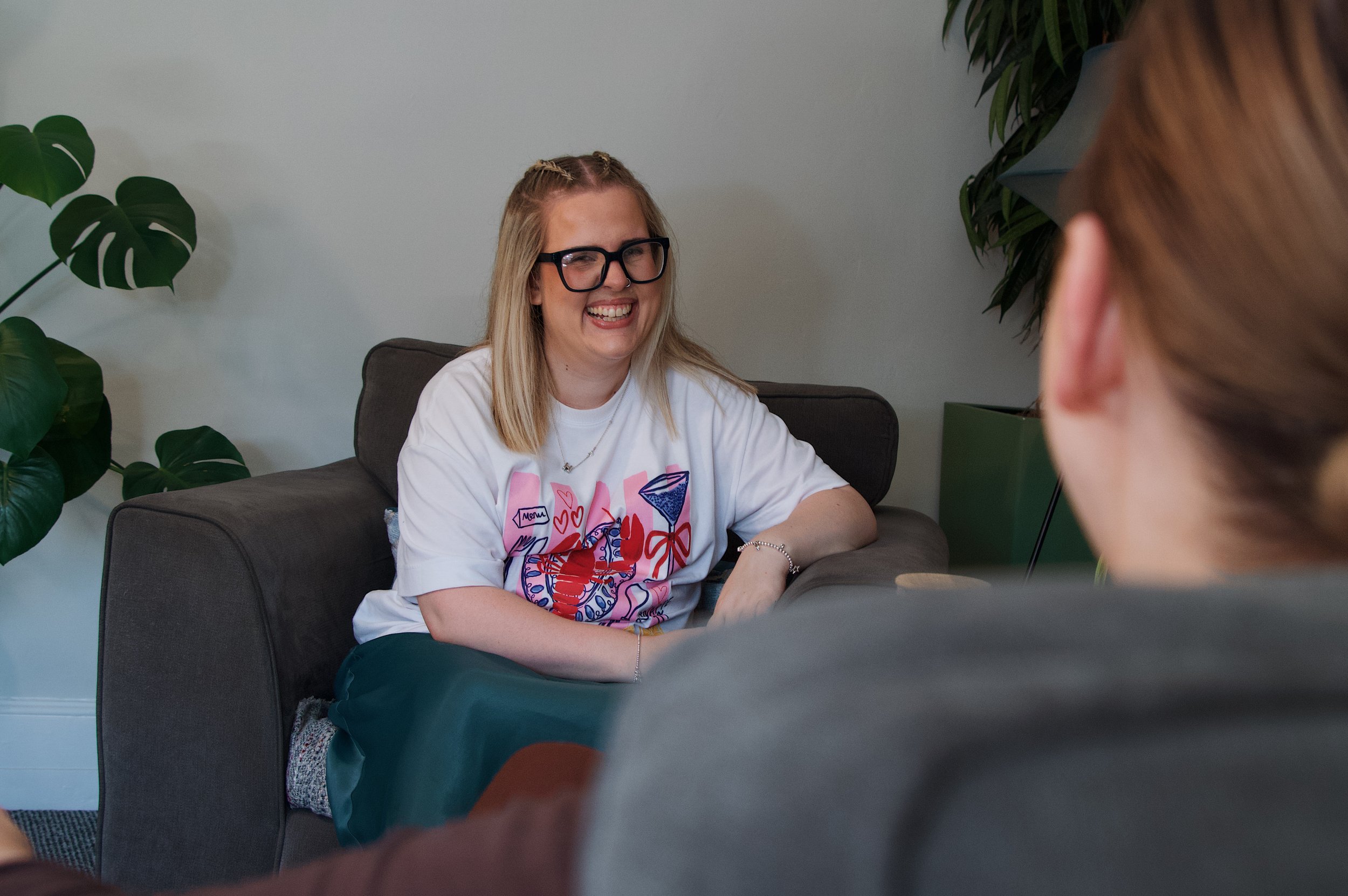 A woman with blonde hair, glasses, and a white shirt smiling during a conversation in a room with green plants.