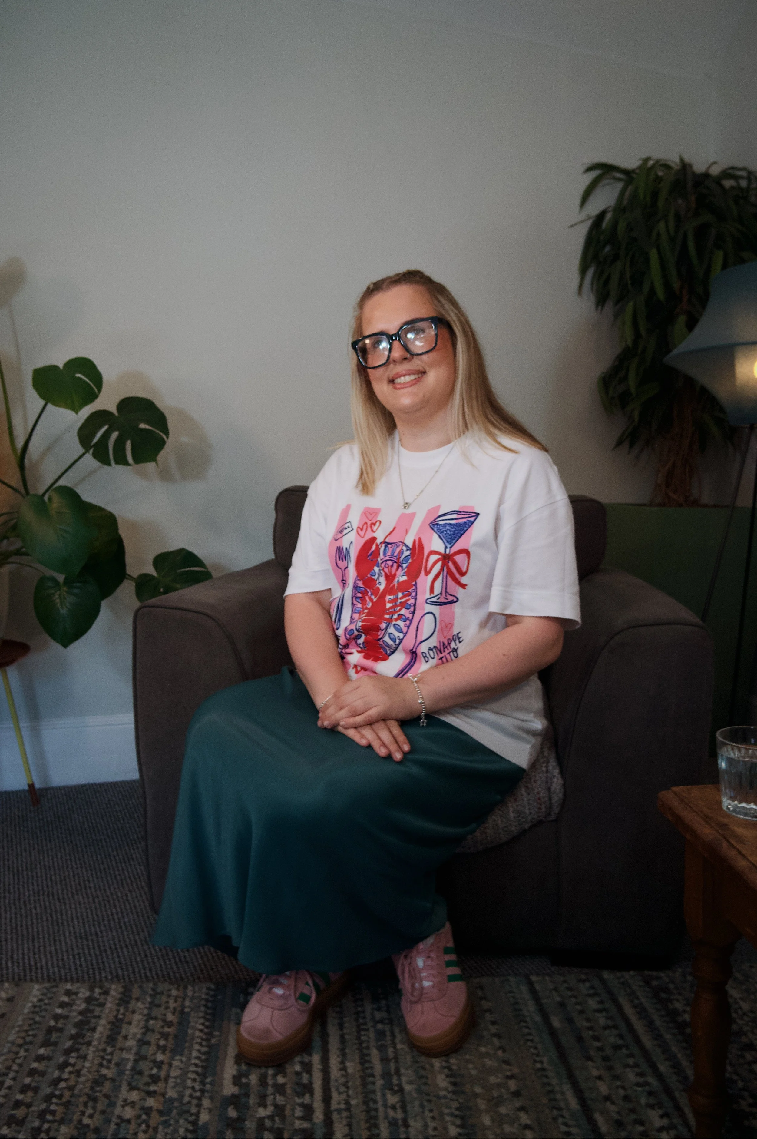 A young woman with blonde hair, wearing glasses, a white graphic t-shirt, and a long green skirt, sitting on a dark couch in a cozy living room. She is smiling and sitting with her hands together on her lap. There are green houseplants, a lampshade, and a small wooden table with a glass of water nearby.