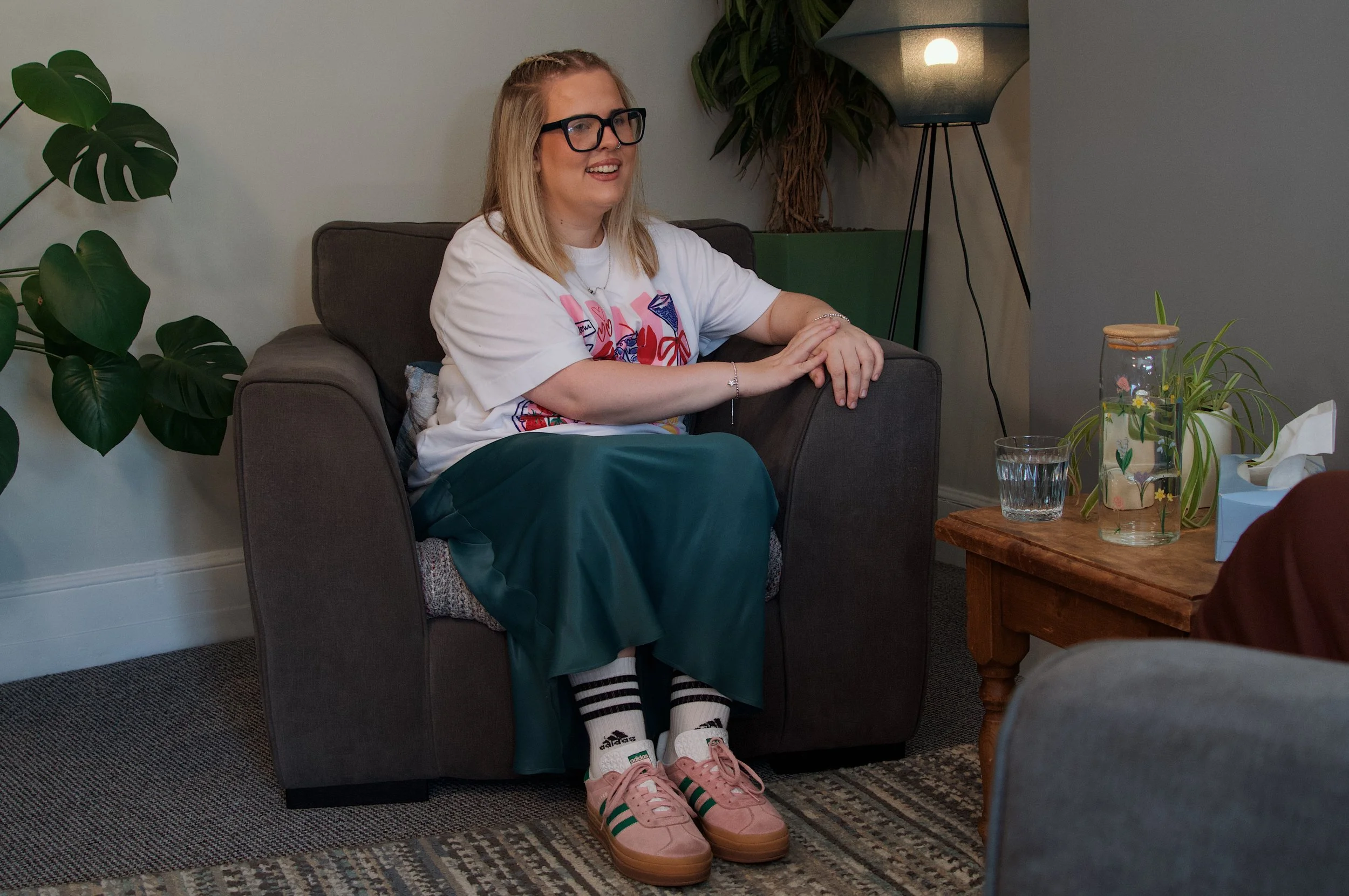 A young woman with blonde hair, glasses, and a bright smile sitting on a dark gray couch in a cozy living room. She is wearing a white T-shirt with colorful graphics, dark green pants, and pink Adidas sneakers. Her legs are crossed, and she is sitting near a wood side table with a glass of water and decorative plants. There is a large leafy houseplant on the left, and a tall blue lamp in the background.