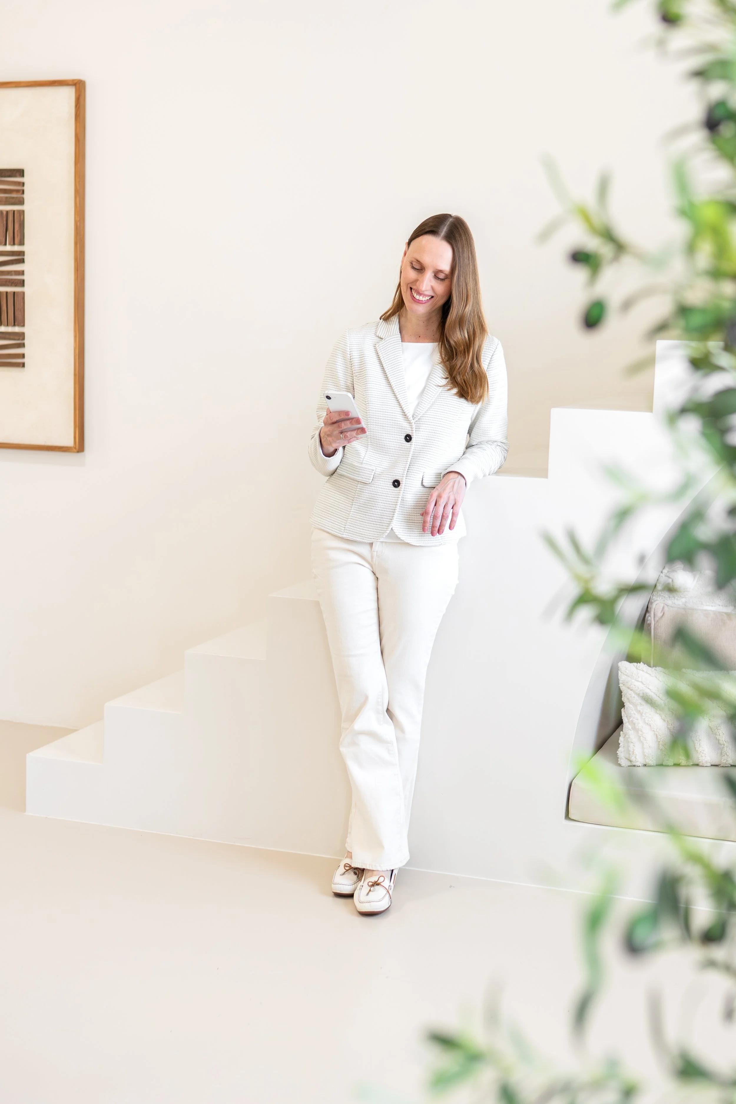 A woman with long brown hair smiling and looking at her phone, dressed in white blazer and pants, leaning against a white wall in a modern, minimalistic room.