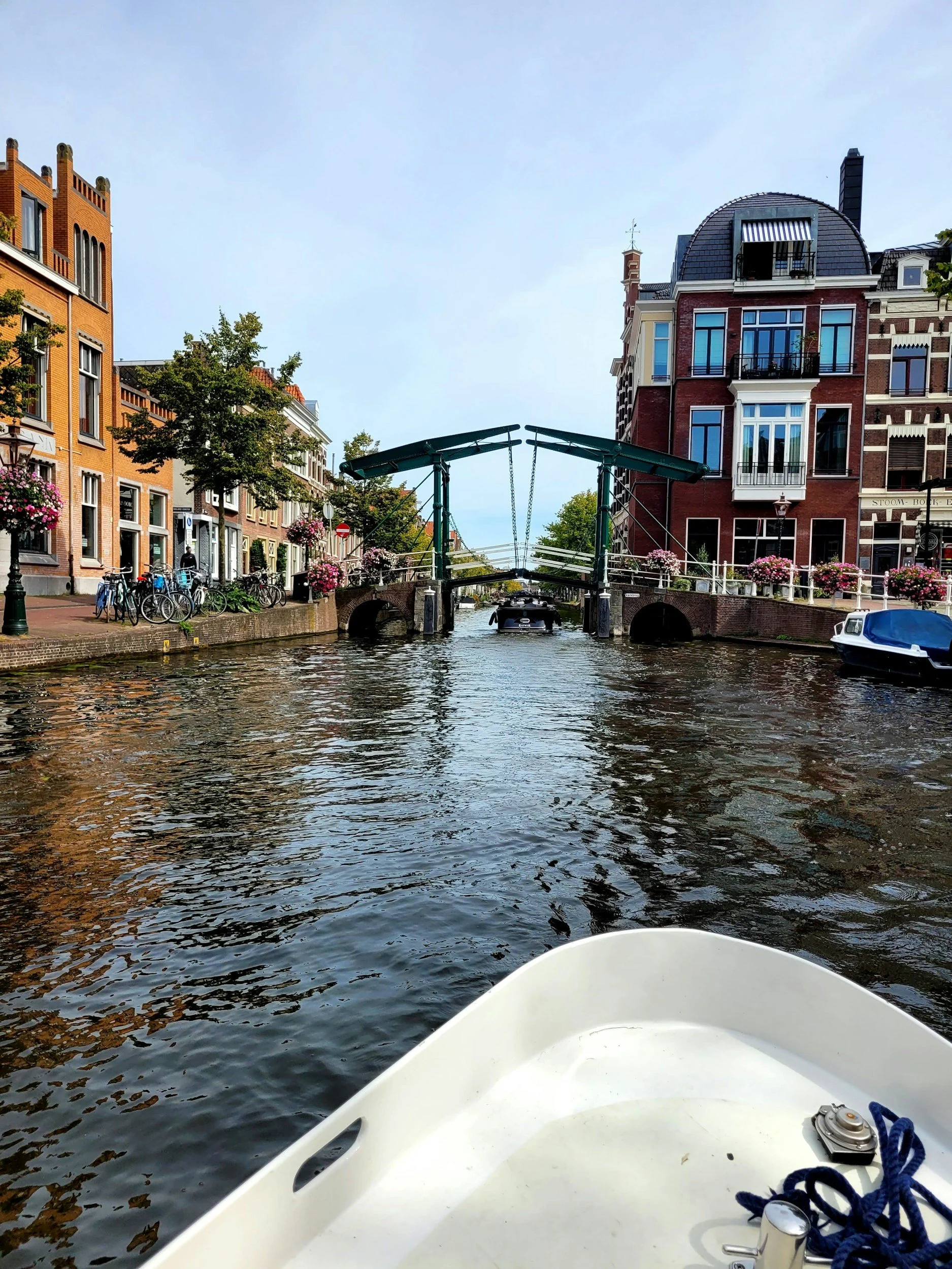 The photo shows a canal scene in a European city, with a white boat in the foreground. The canal is lined with colorful, historic buildings, trees, and a small bridge. A boat is passing under the bridge, and bicycles are parked along the street.