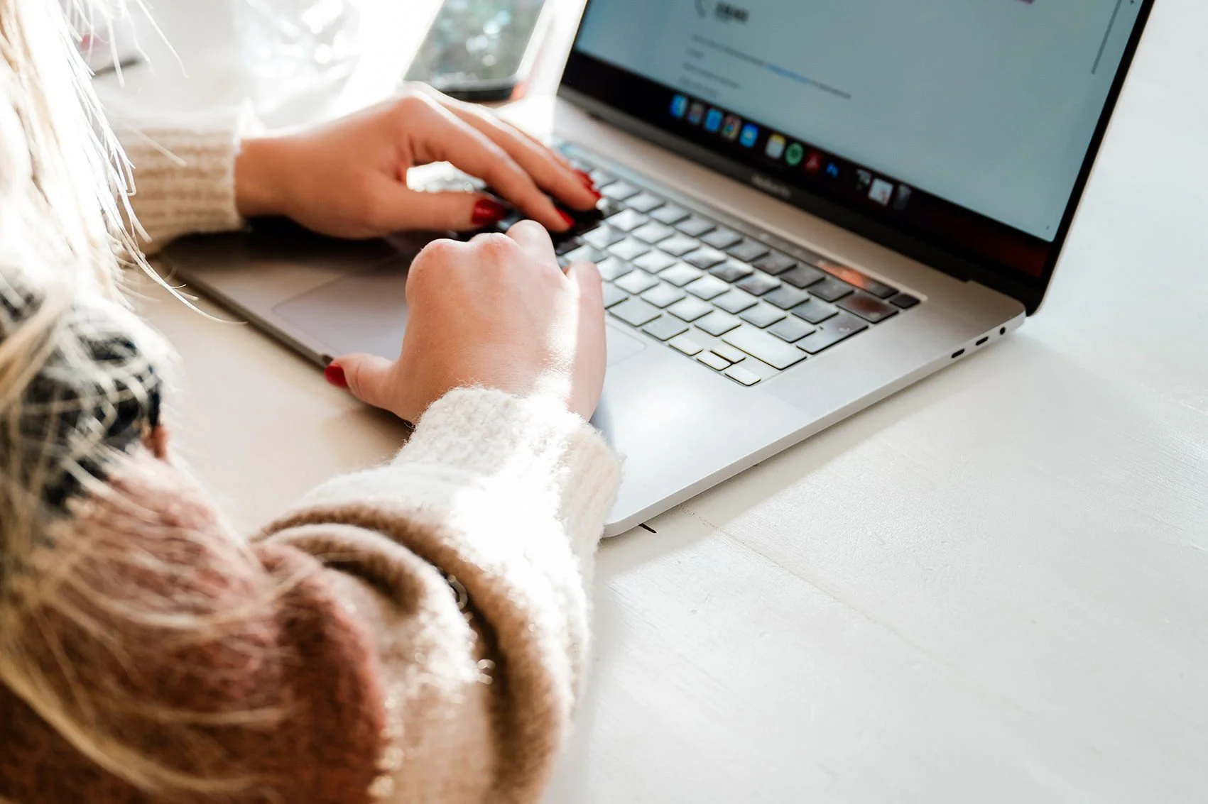 A person typing on a silver laptop with a white keyboard, sitting on a light-colored desk.