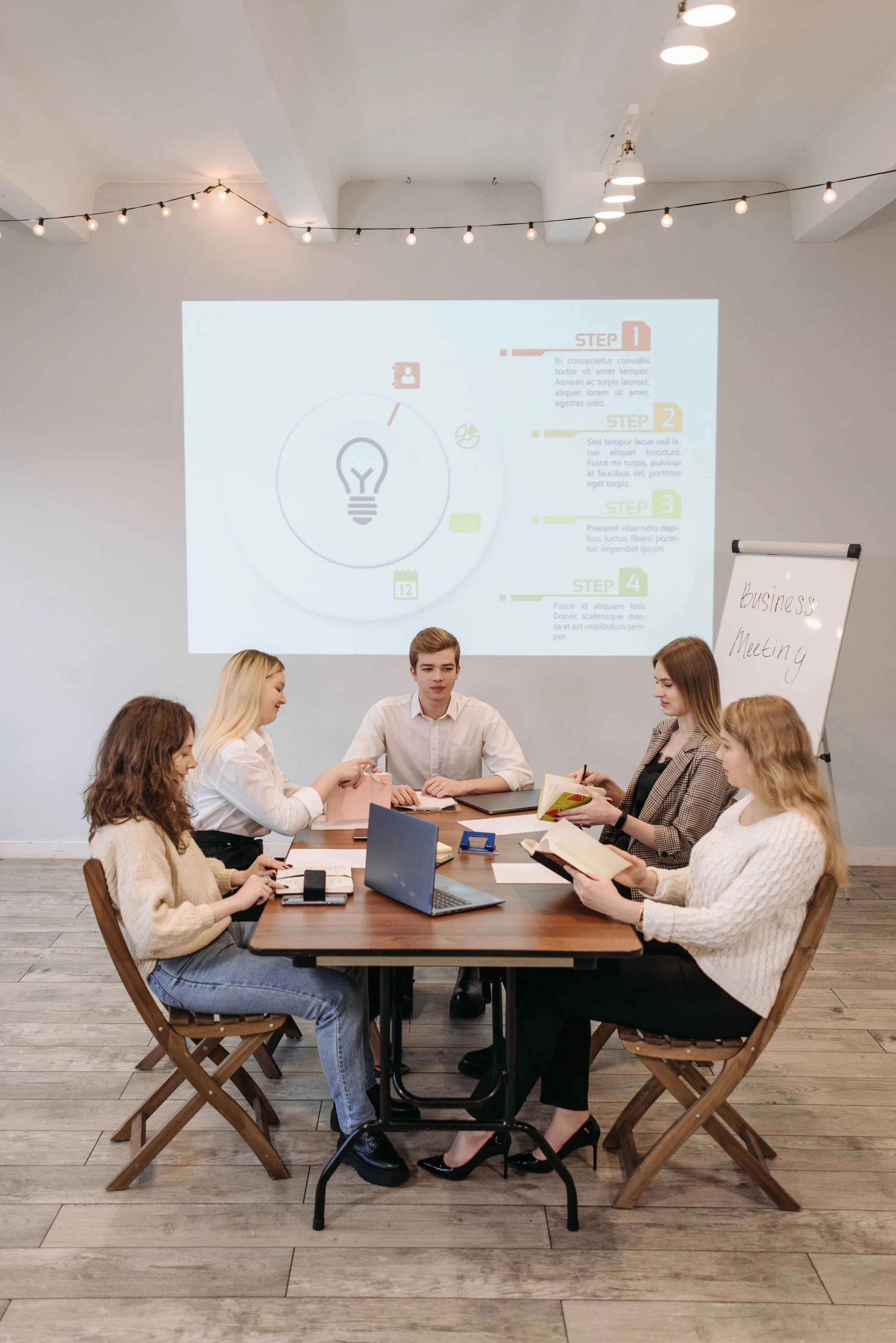 A group of seven young adults participating in a business meeting or workshop around a wooden table in a modern, well-lit room with a white wall and a projector screen showing a presentation.