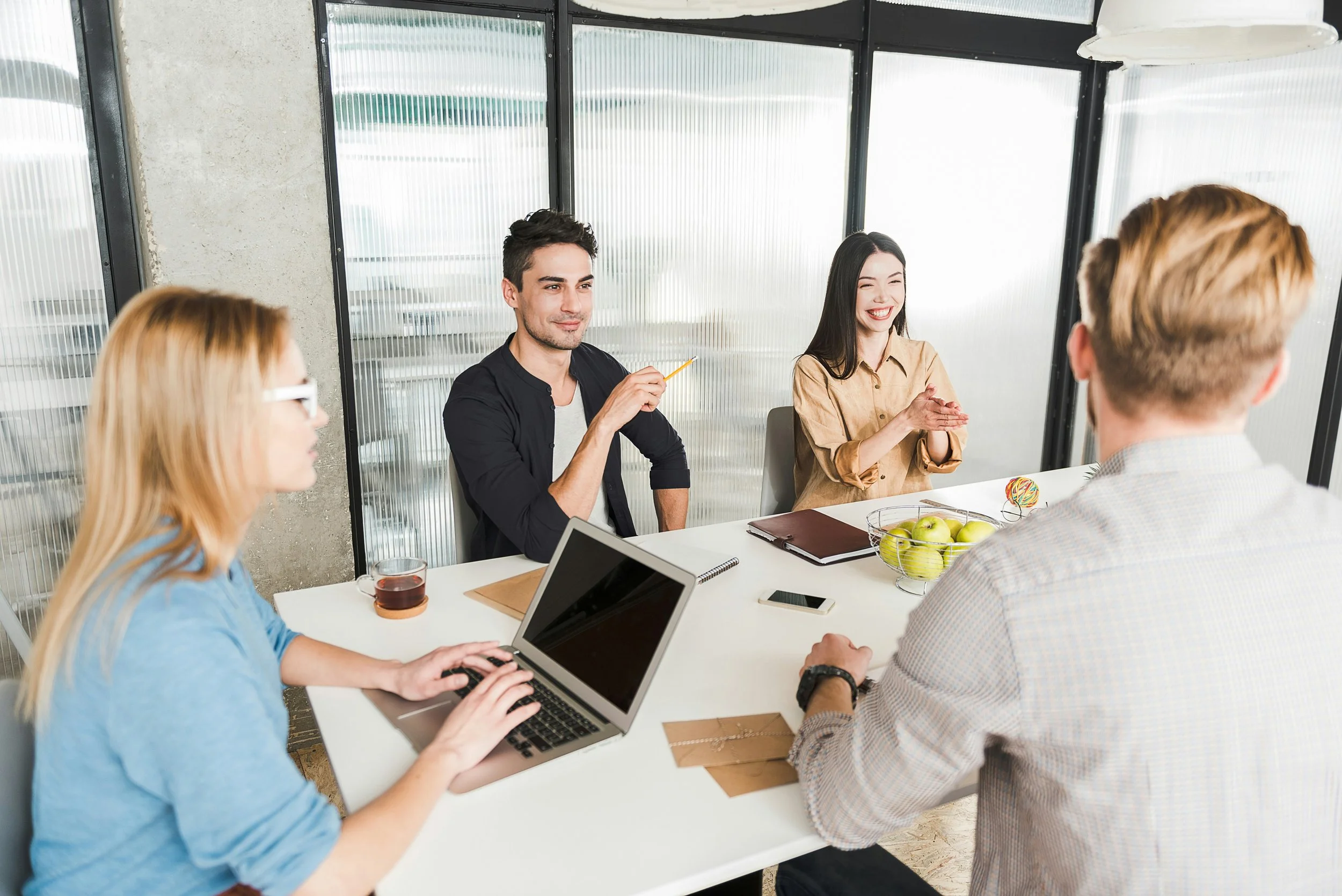A group of five people gathered around a white table in a modern office, engaged in a discussion. Two women are using laptops, one with a tablet in hand, and a man holding a pencil and smiling. The table has notebooks, a phone, and a fruit bowl with apples, with a coffee mug and a drink also visible. They are seated near large windows with frosted glass panels, and a large hanging lamp is above the table.