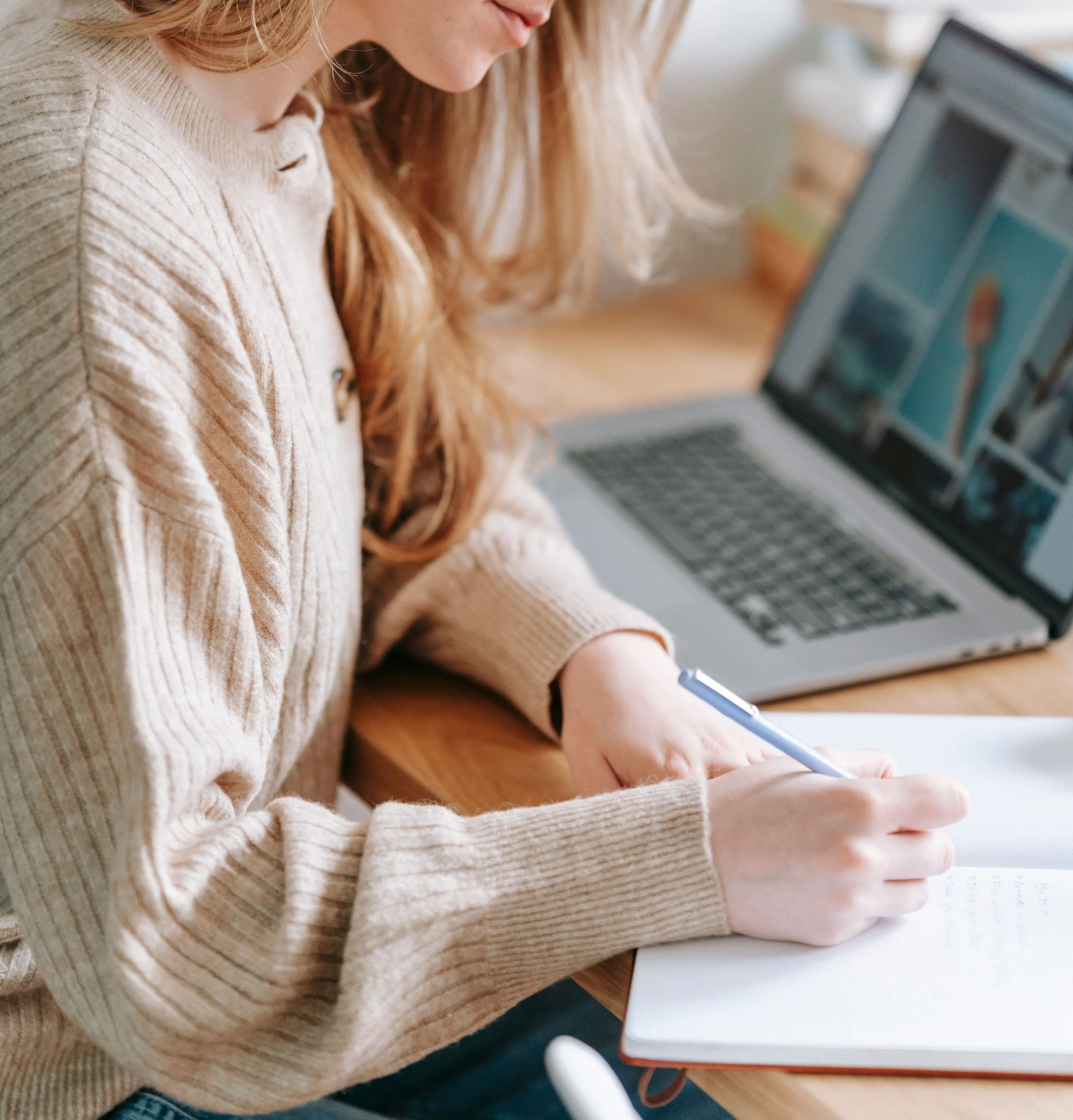 Close-up of a woman writing in a notebook with a pen, sitting at a desk with a laptop nearby.