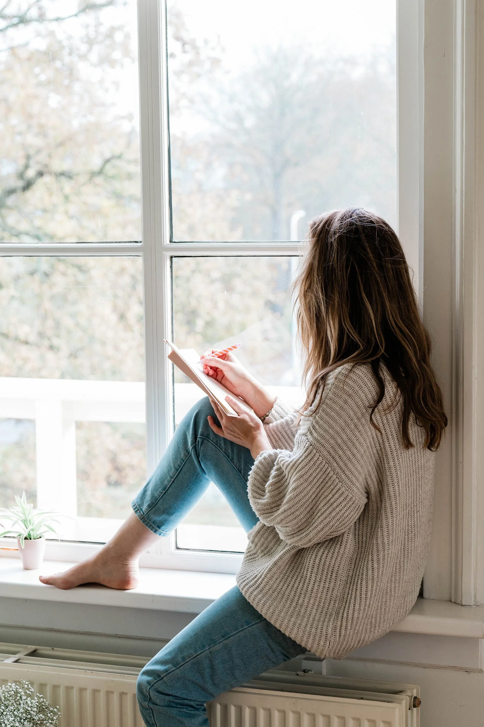 A young woman with long hair in a beige sweater and blue jeans sitting on a window sill, reading a book, with her feet resting on a radiator, next to a potted plant and looking out the window.