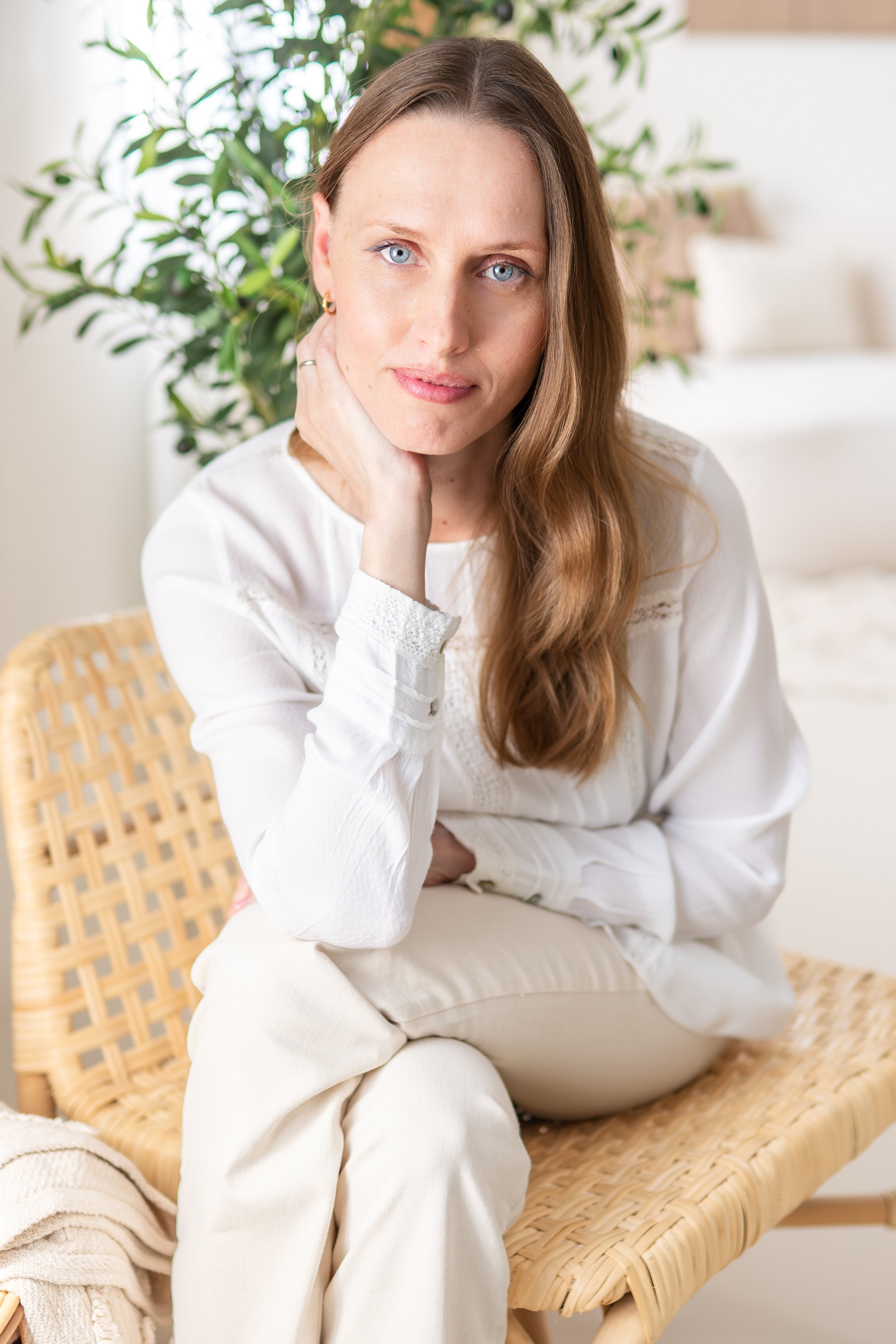 A woman with long brown hair and blue eyes sitting on a wicker chair in a bright room with green plants in the background. She is wearing a white long-sleeve top and light-colored pants, resting her hand on her neck and looking at the camera.