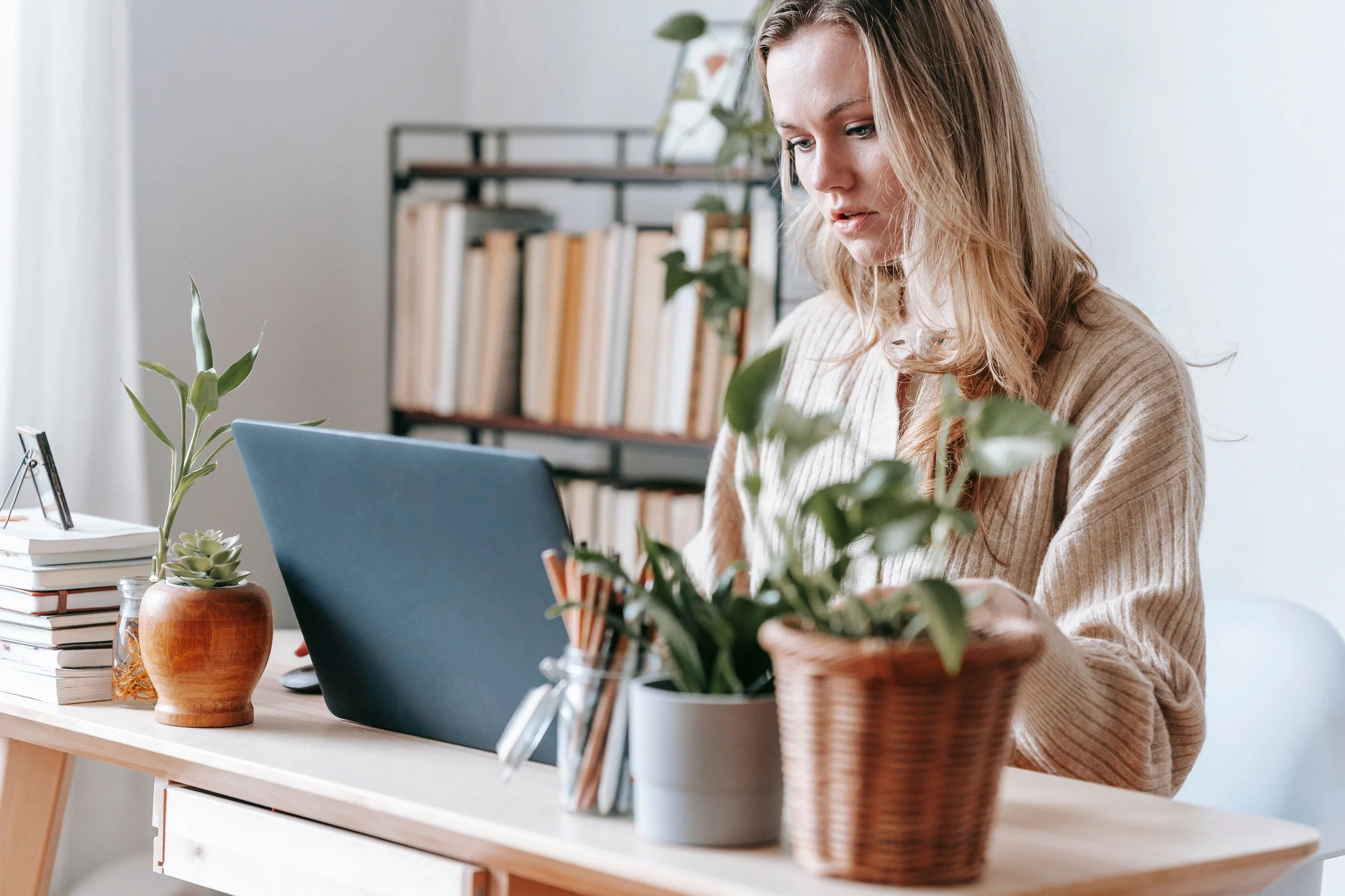 Woman working at a desk with plants, books, and a laptop in a bright, organized home office.